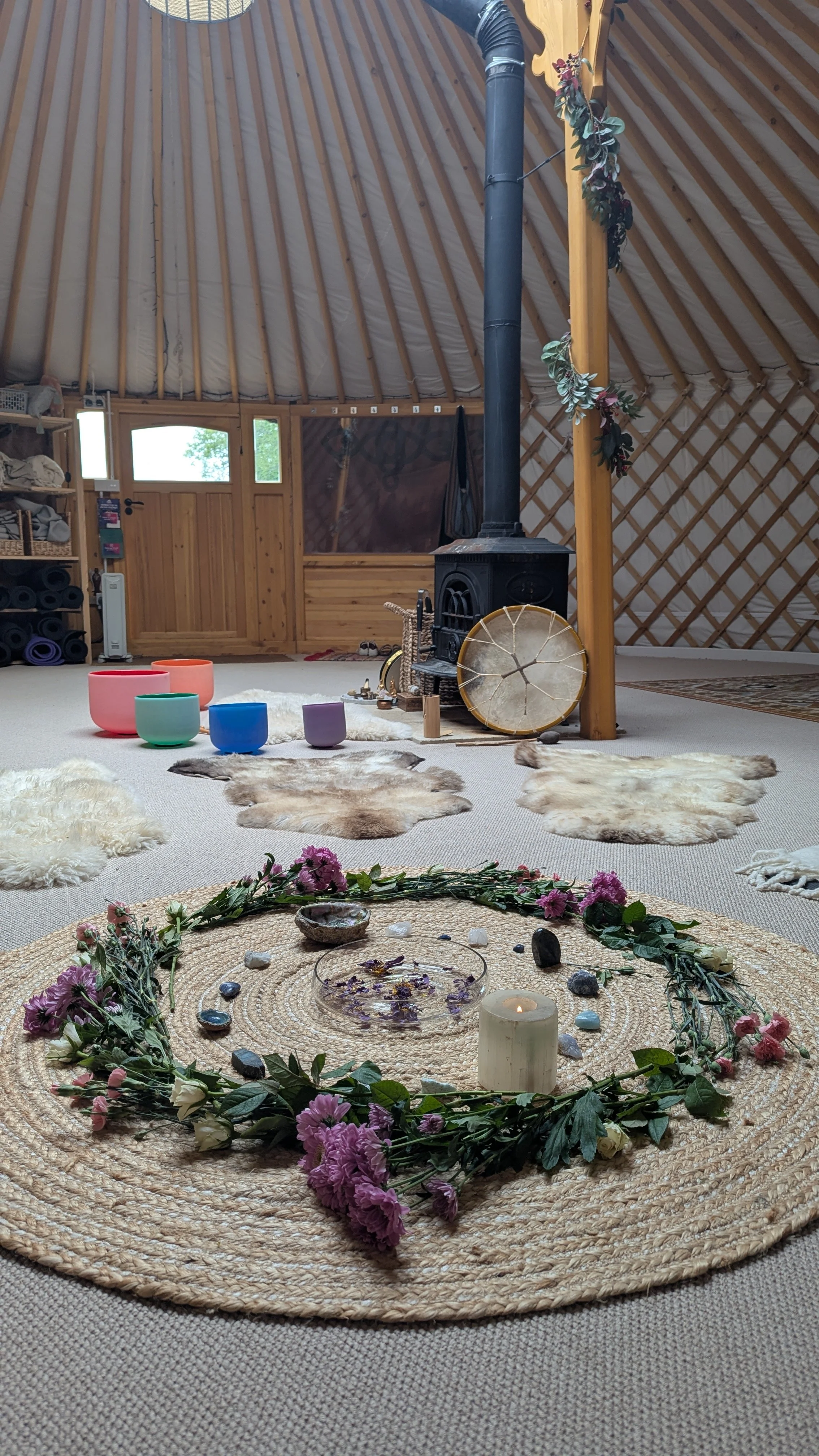A cozy yurt interior with a central circular woven rug, candles, flowers, and crystals, surrounded by animal pelts, with a wood stove, colorful singing bowls, and shelves with yoga mats in the background.