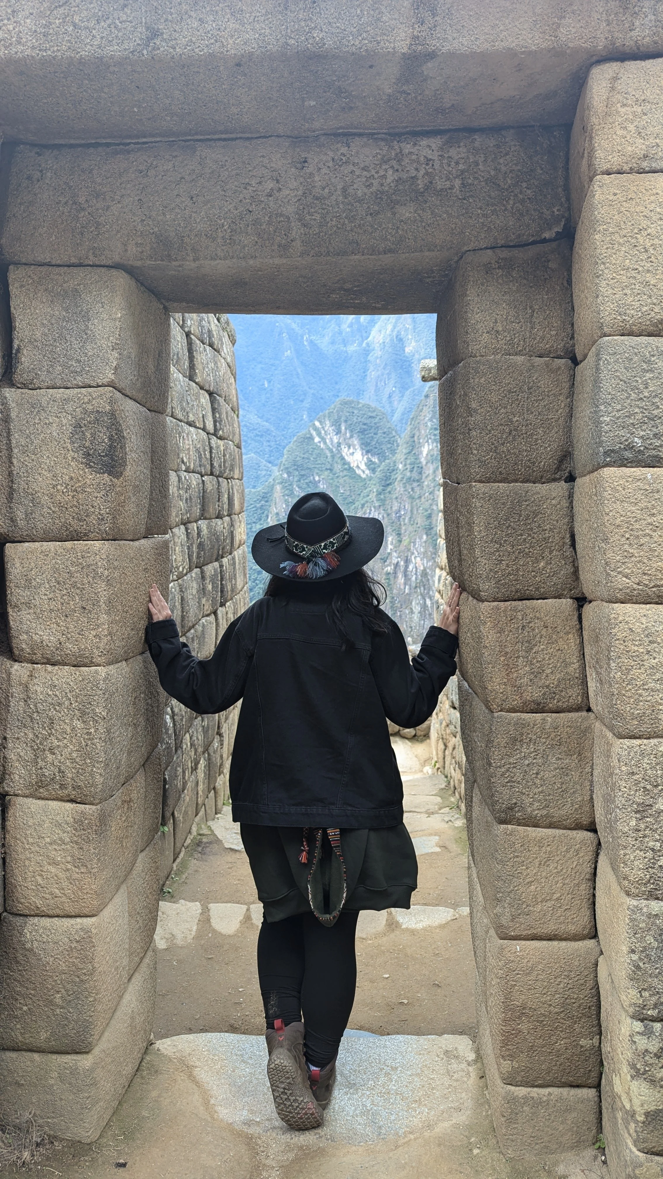 A woman wearing a black hat and black jacket walking through an ancient stone doorway at Machu Picchu, with lush green mountains in the background.