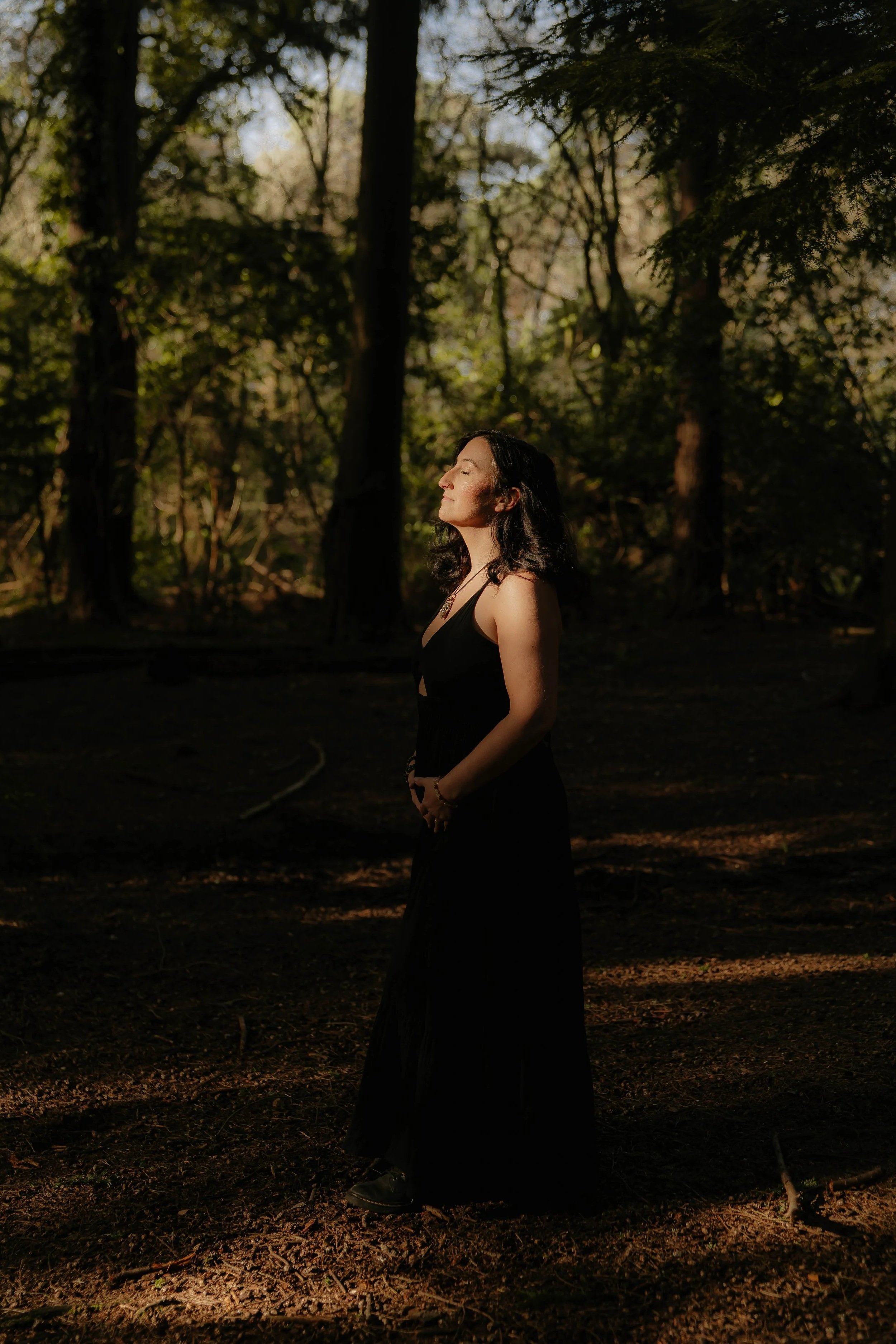 A woman with dark hair wearing a black dress stands in a forest with her eyes closed, illuminated by sunlight filtering through the trees.