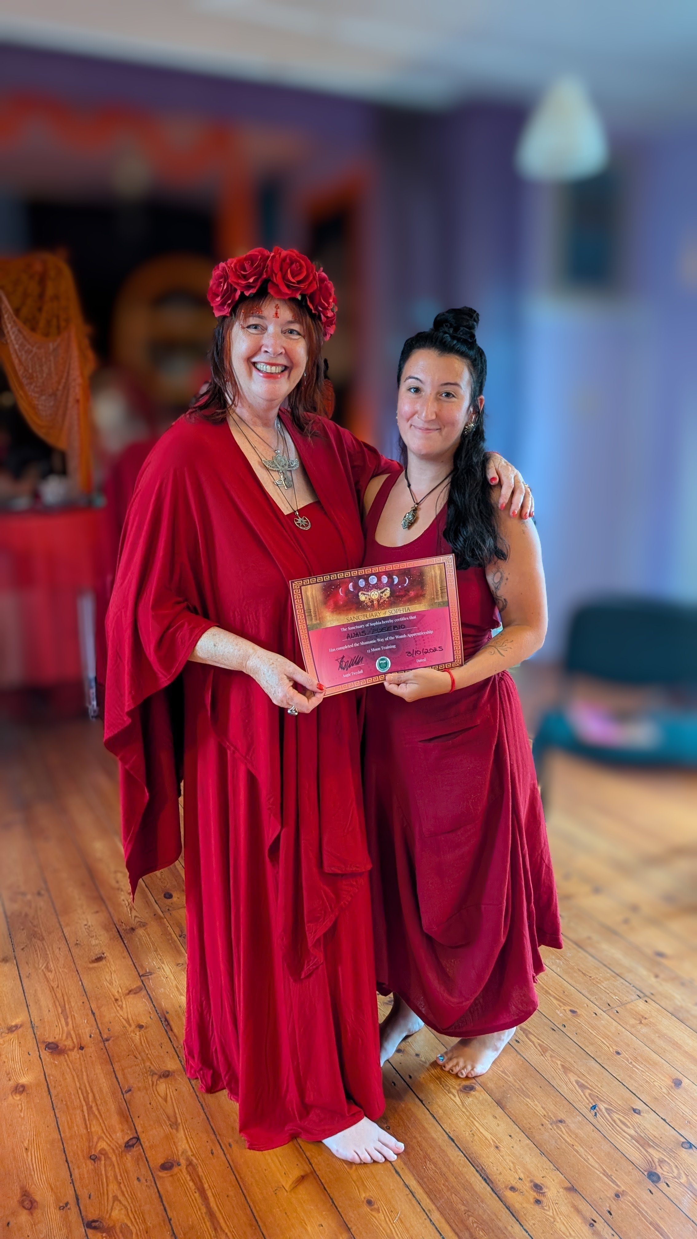 Two women dressed in red, one wearing a flower crown, smiling, while holding a certificate, in a decorated indoor space with wooden floors.