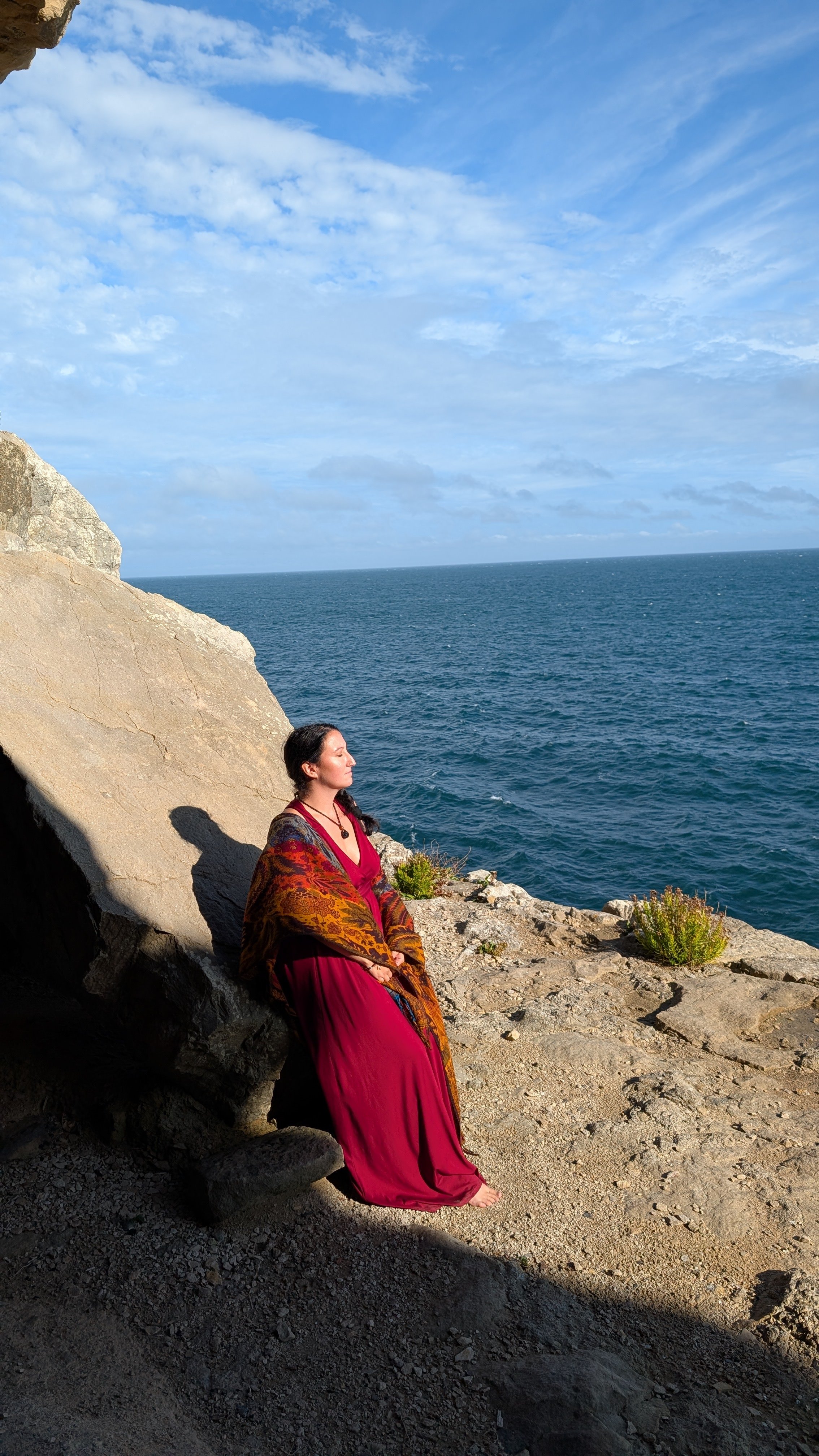 Woman in a red dress sitting on rocks by the ocean, with her eyes closed and the sky partly cloudy.