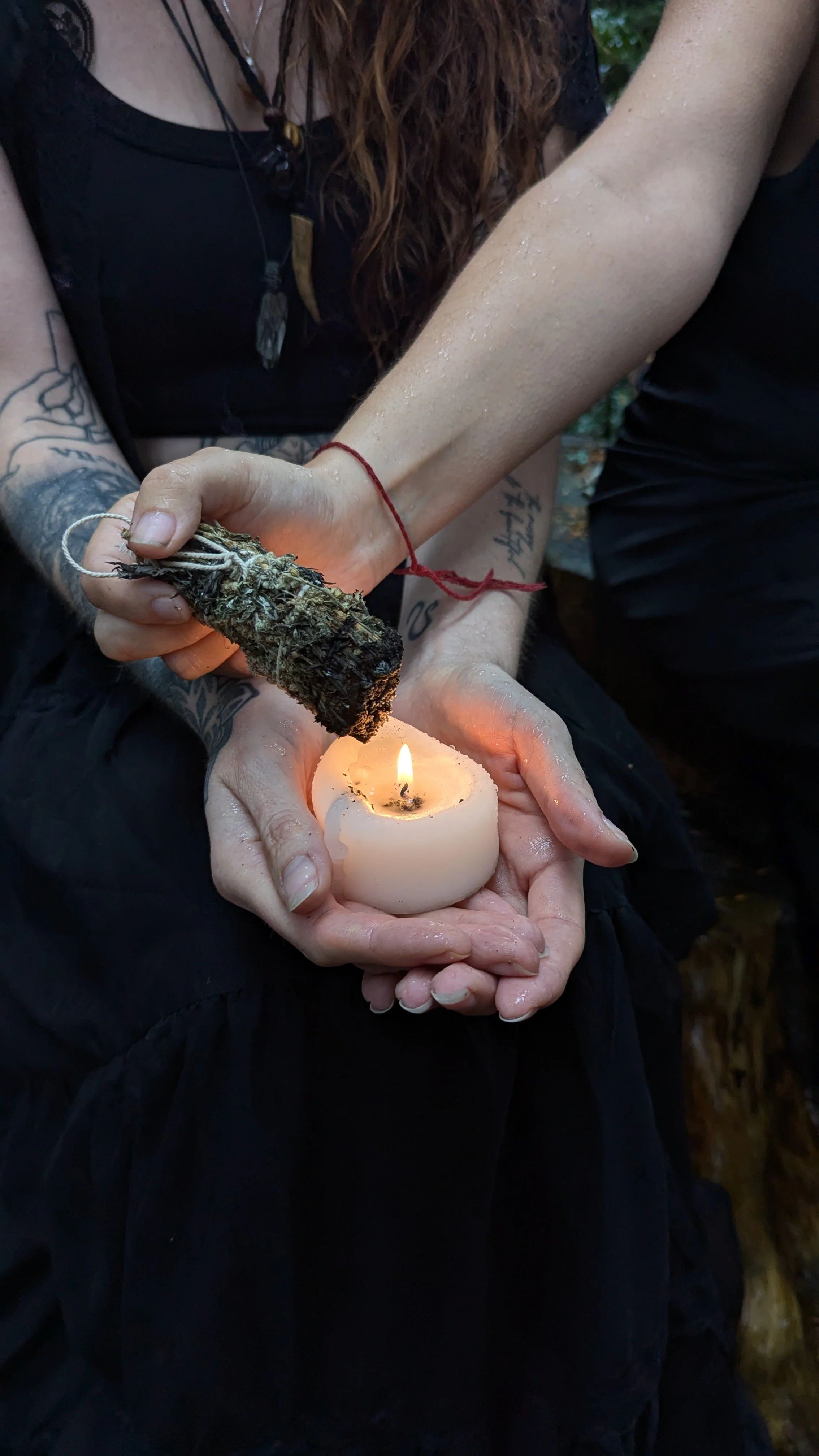 Two people are holding a candle and a small bundle of herbs or plant material with a fire source. The person on the left, who has tattoos and tattoos on their arms, is holding the herbs, while the person on the right holds the candle with both hands. The scene appears to be outdoors.