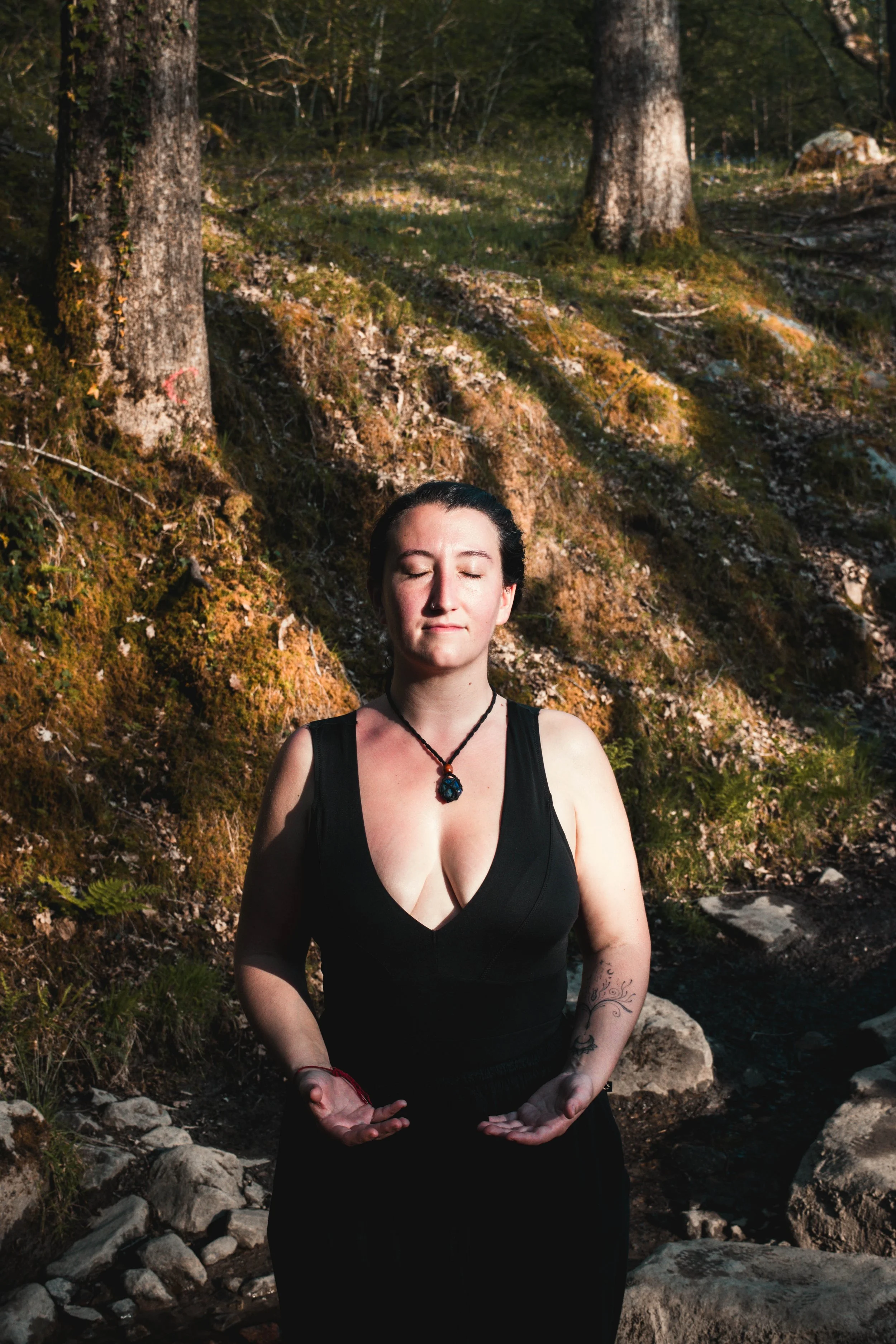 A woman with dark hair in a bun, wearing a black sleeveless dress, stands outdoors in a forested area with tall trees and mossy rocks, with her hands in a meditative pose and eyes closed.