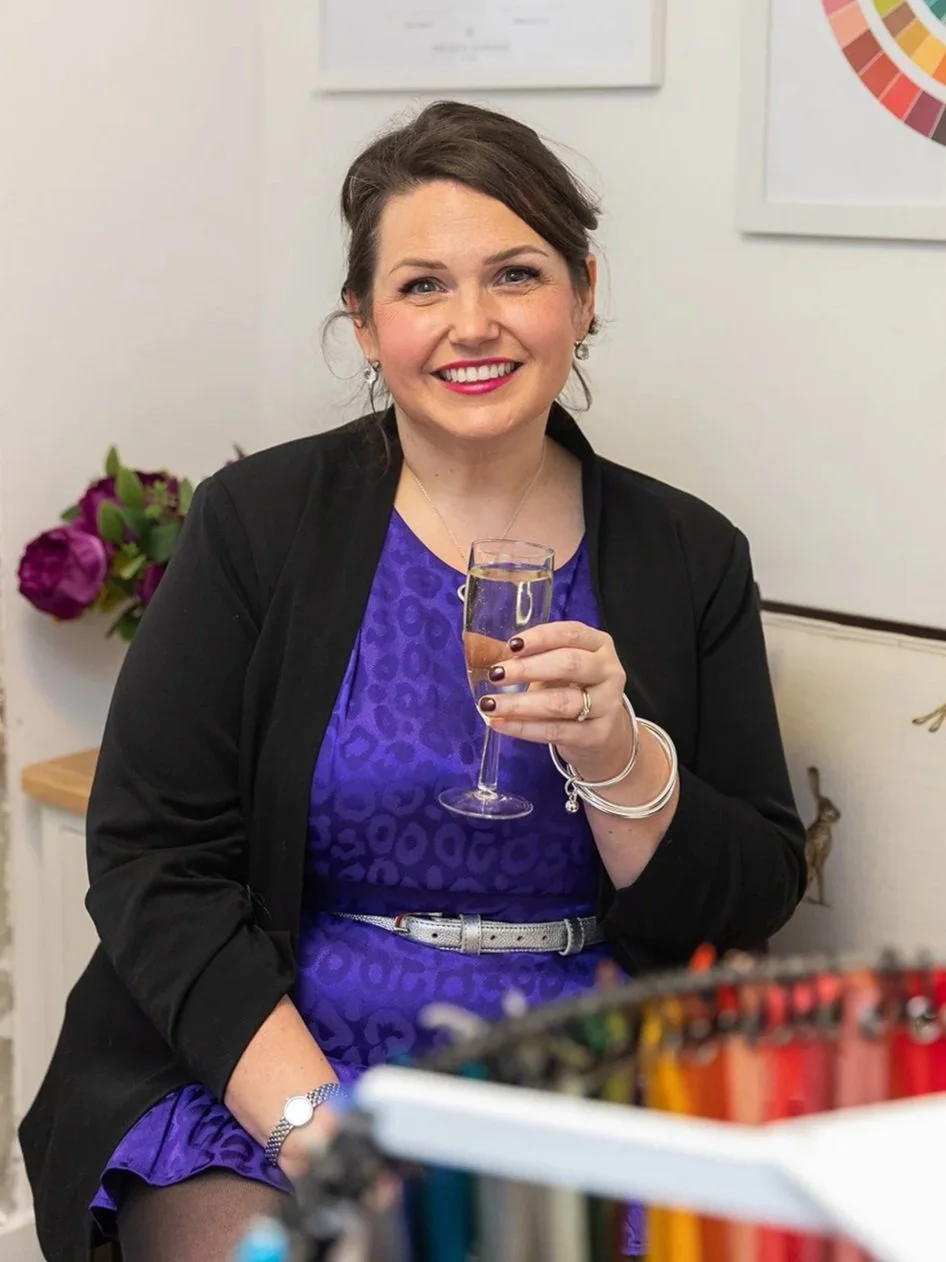 Lucy, a smiling stylist in a vibrant satin purple dress and black blazer holds a glass of fizz in her studio, surrounded by colour analysis drapes and charts, creating a warm and inviting styling space.