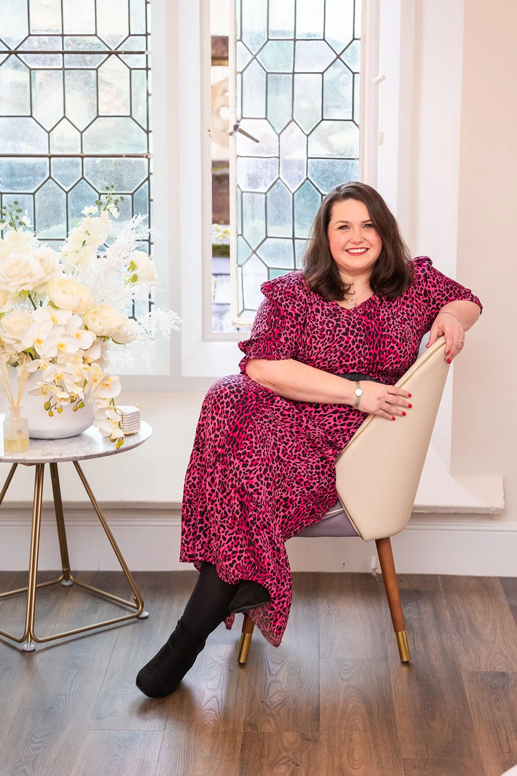 Lucy, a style consultant in a vibrant pink animal-print dress poses confidently in a modern, light-filled space, showcasing bold personal style beside a chic floral arrangement and contemporary décor.