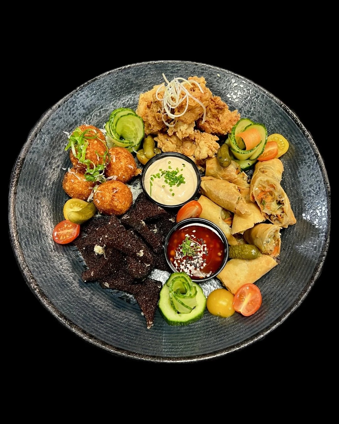 A glass plate with various Asian appetizers, including fried calamari, spring rolls, crispy noodles, purple rice chips, and small bowls of dipping sauces, garnished with vegetables and herbs.