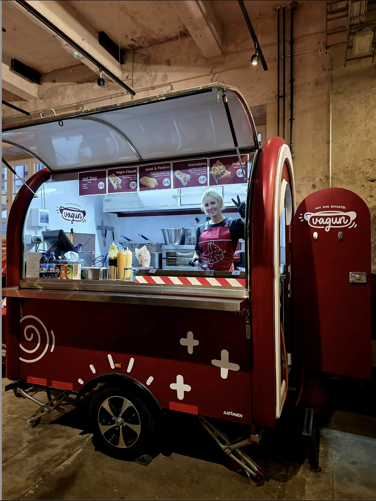 A woman smiling and making a peace sign inside a vintage red mobile food stand that sells hot dogs and other fast food, with a menu board above her.