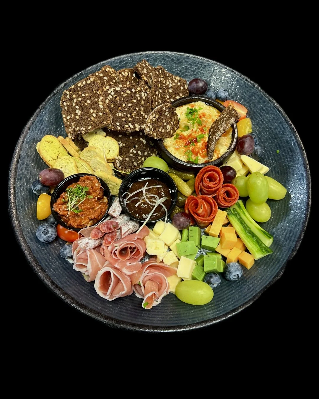 A charcuterie and cheese platter with crackers, sliced meats, cheeses, grapes, blueberries, cherry tomatoes, cucumber slices, dips, and crackers on a black plate.