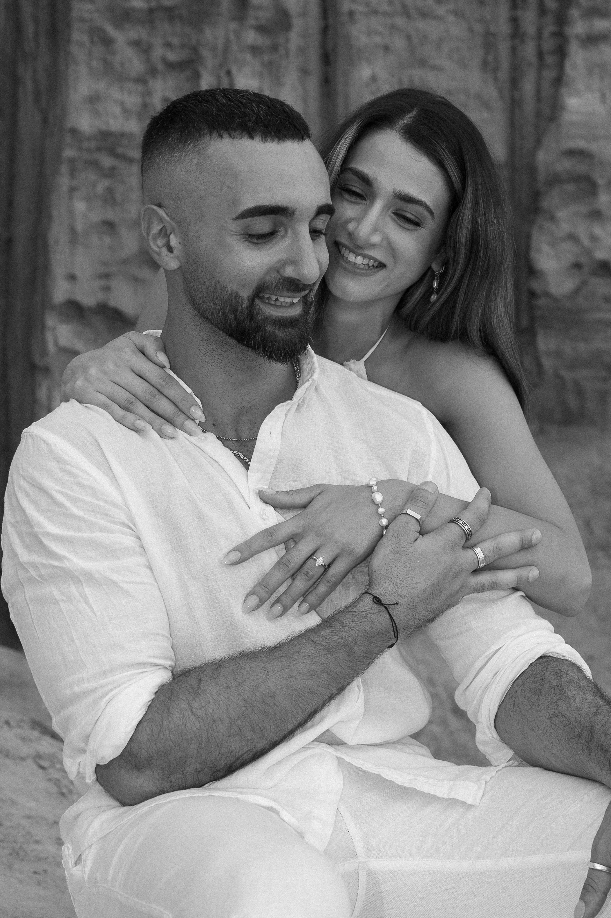 A happy couple sitting close together, smiling and showing their rings, with the woman embracing the man from behind, against a rustic stone wall background.