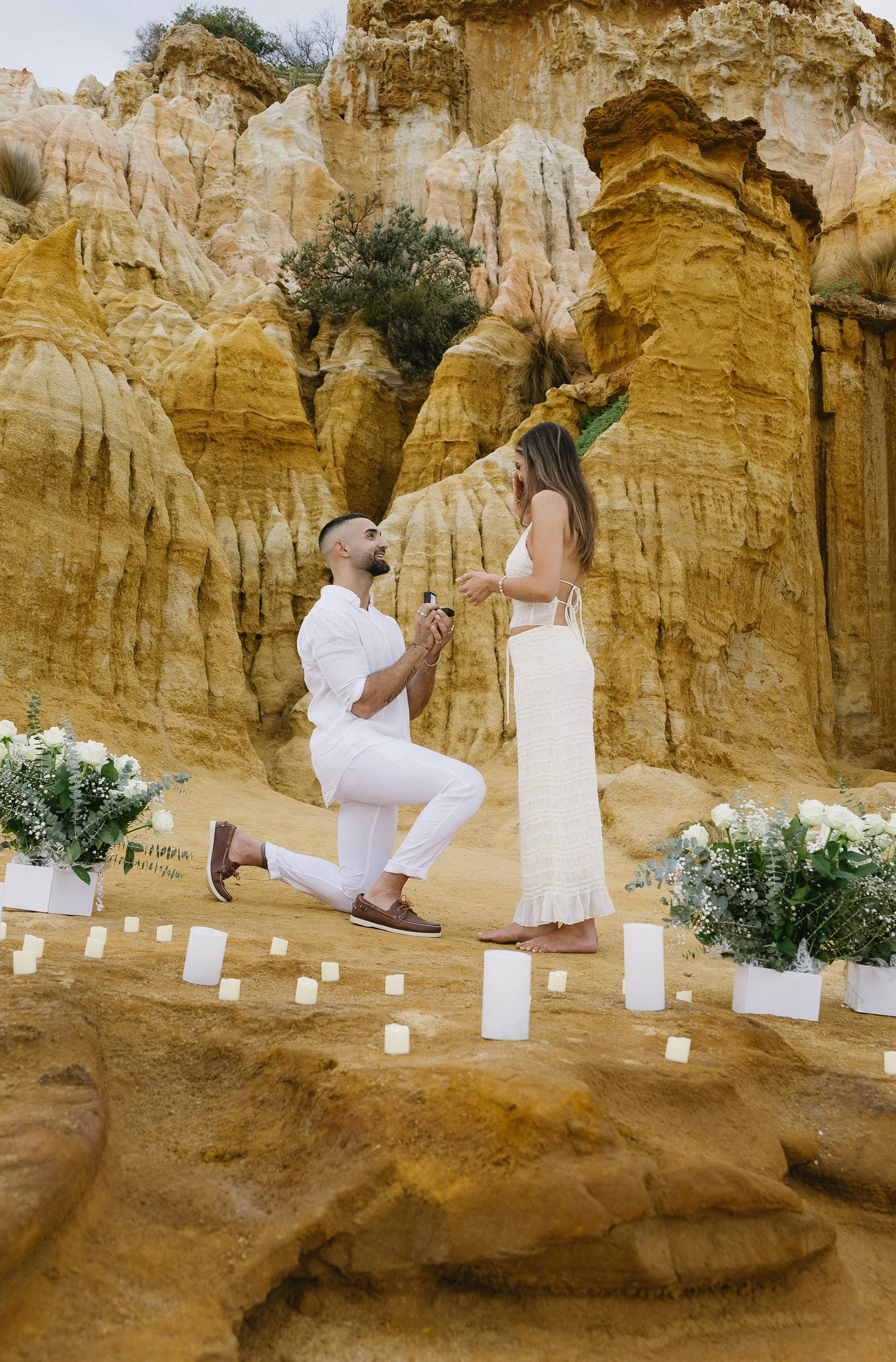 A man proposing marriage to a woman in a desert-like setting with orange and yellow rocky formations, surrounded by flowers and candles.
