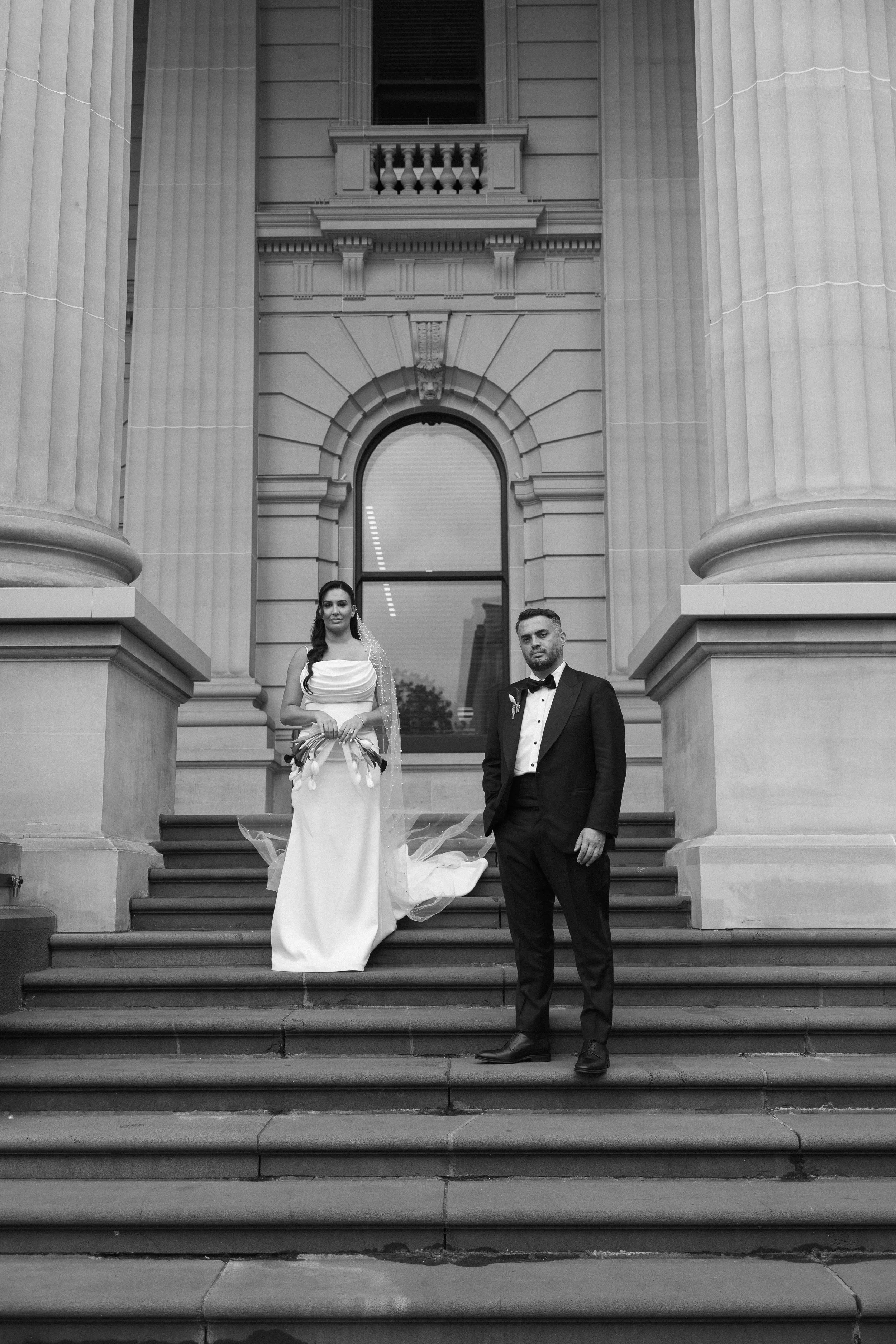 Black and white photo of a bride and groom standing on steps outside a classical building with large columns and an arched window.