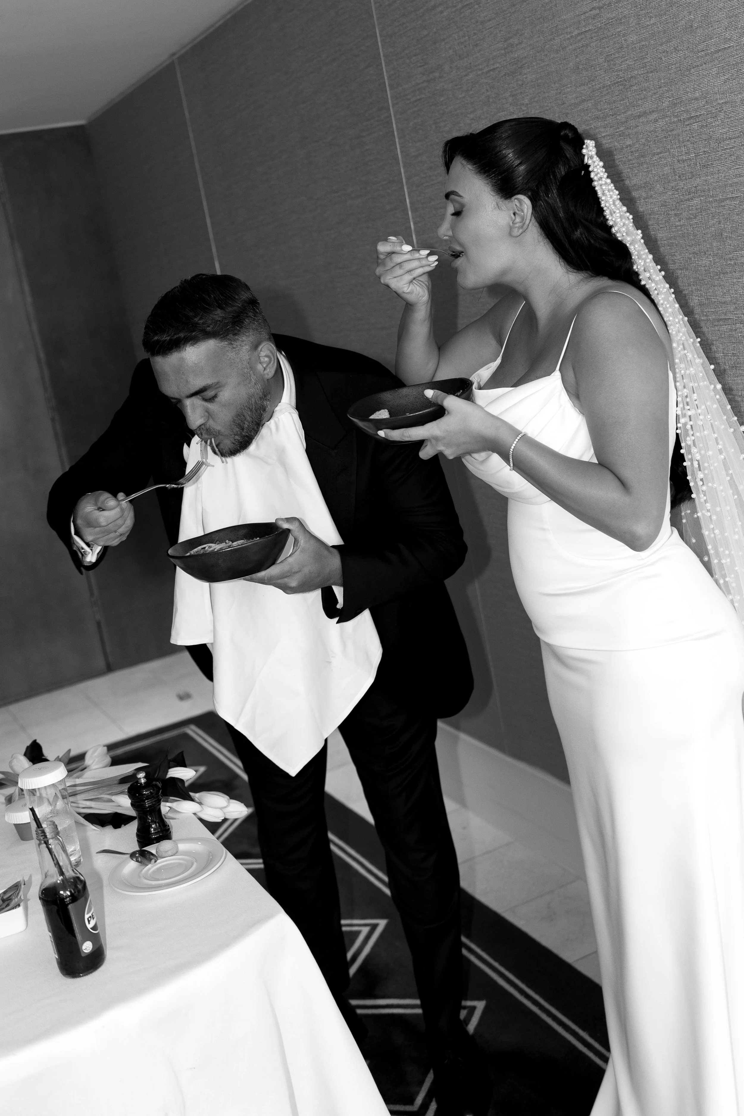 A bride in a white wedding dress and veil eats from a bowl while a groom in a tuxedo with a bib eats from a bowl, sharing a moment during their wedding celebration.