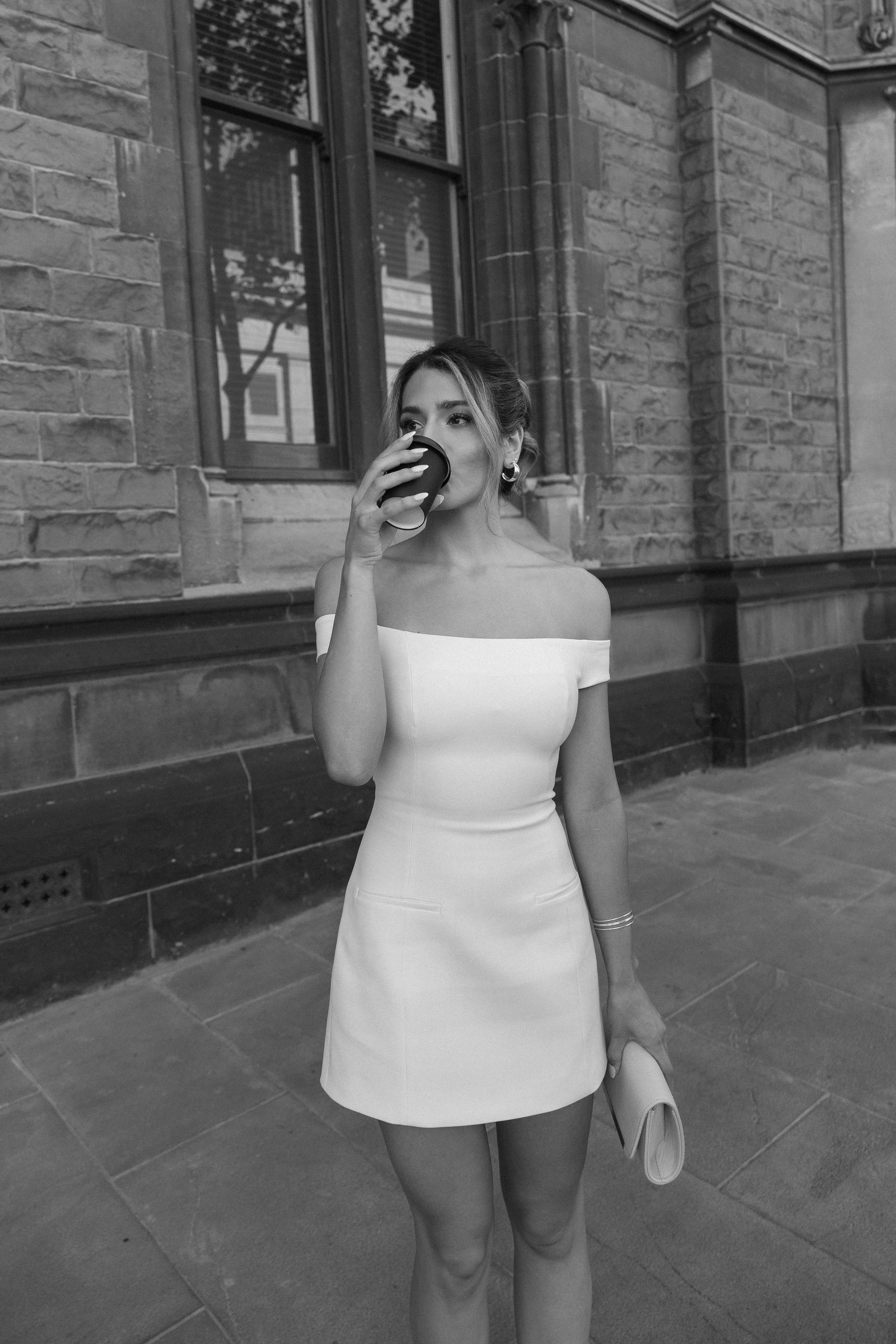 A woman in an off-shoulder dress drinks from a takeaway coffee cup on sidewalk outside a brick building.