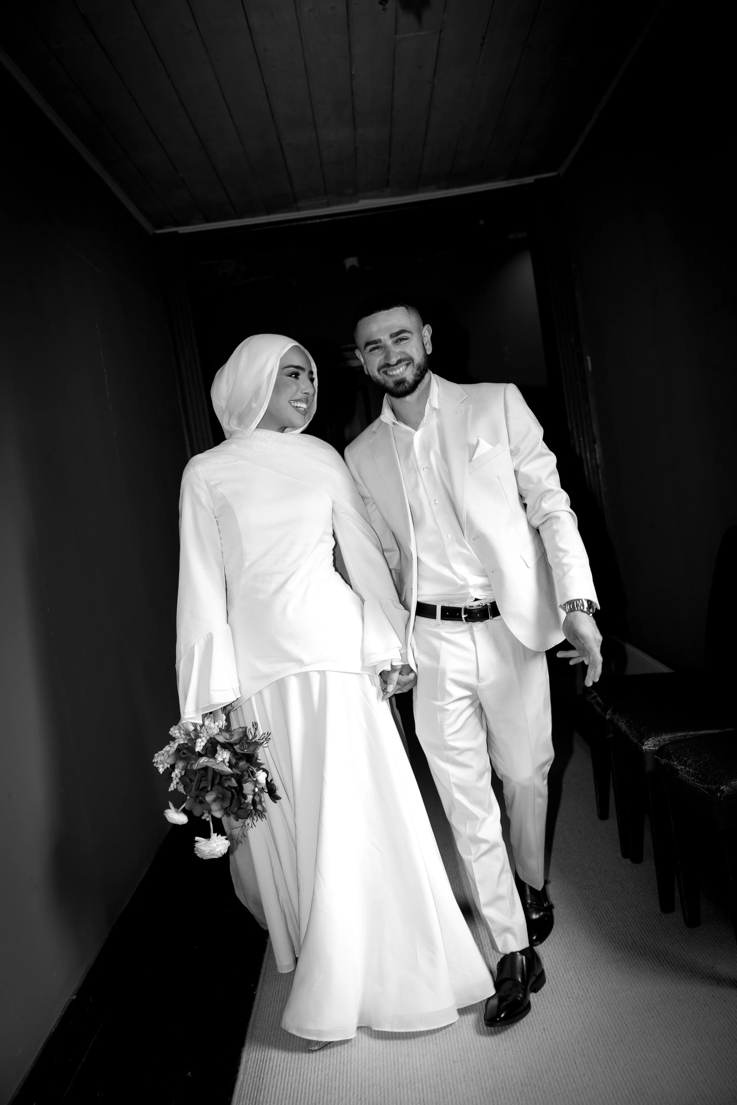 A bride and groom walking together, holding hands, in a hallway. The bride is wearing a modest white dress and headscarf, holding a bouquet of flowers. The groom is wearing a white suit jacket, shirt, and dark shoes. Both are smiling and looking happy.