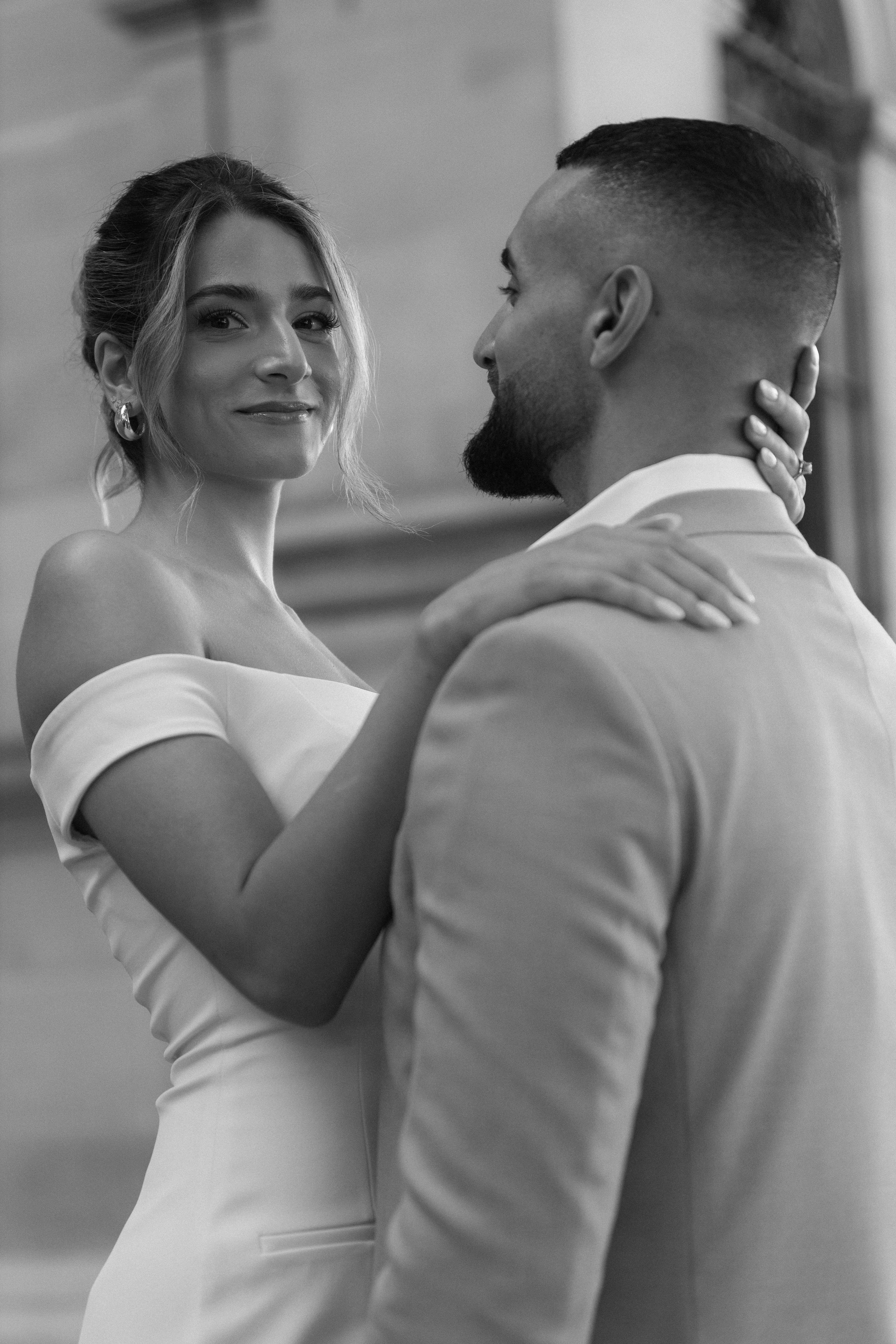 A woman in a white off-shoulder dress and a man in a suit dance closely, smiling, in a black and white photo.