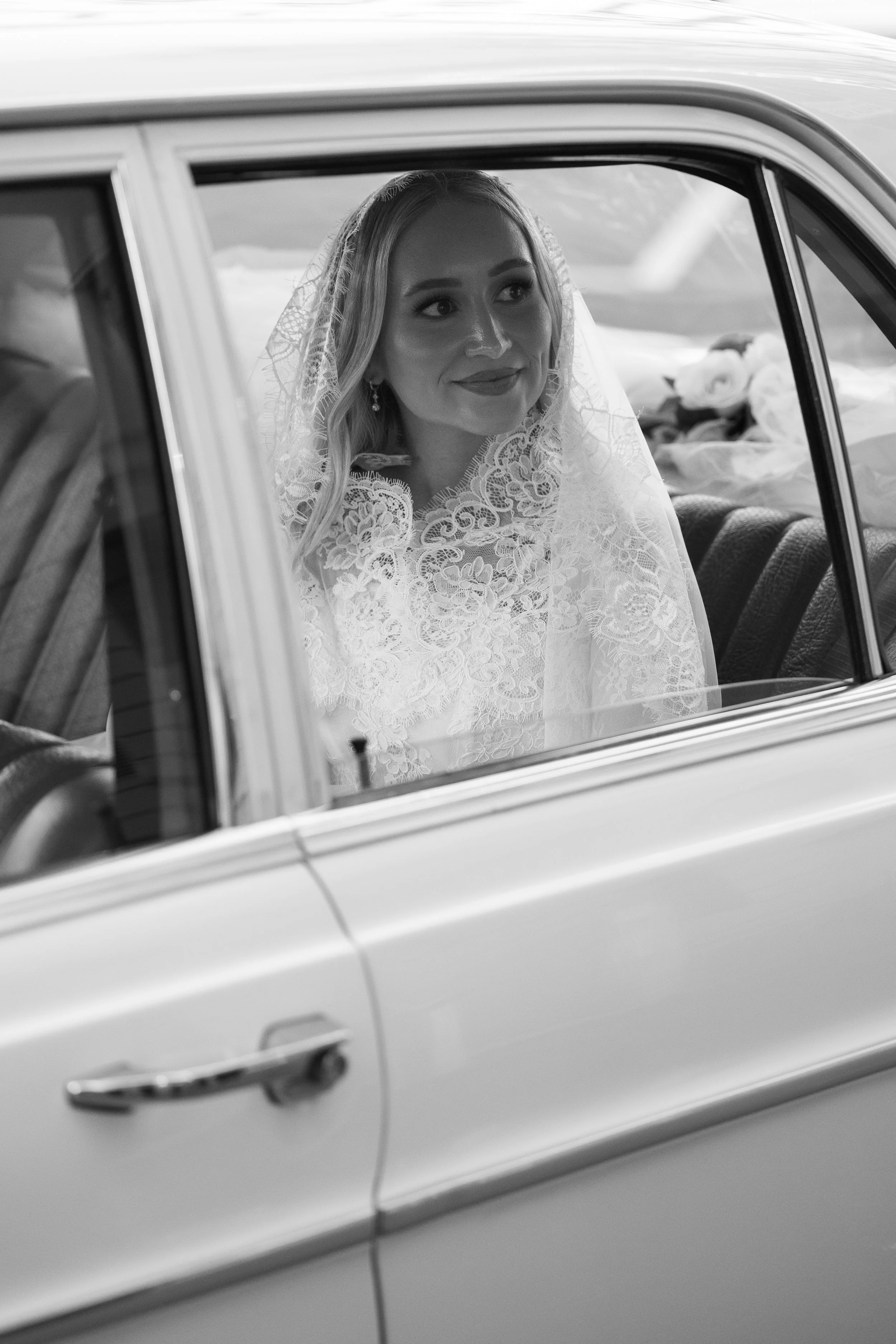 A bride smiling inside a vintage car, wearing lace wedding dress and veil, holding bouquet, looking out the open window.