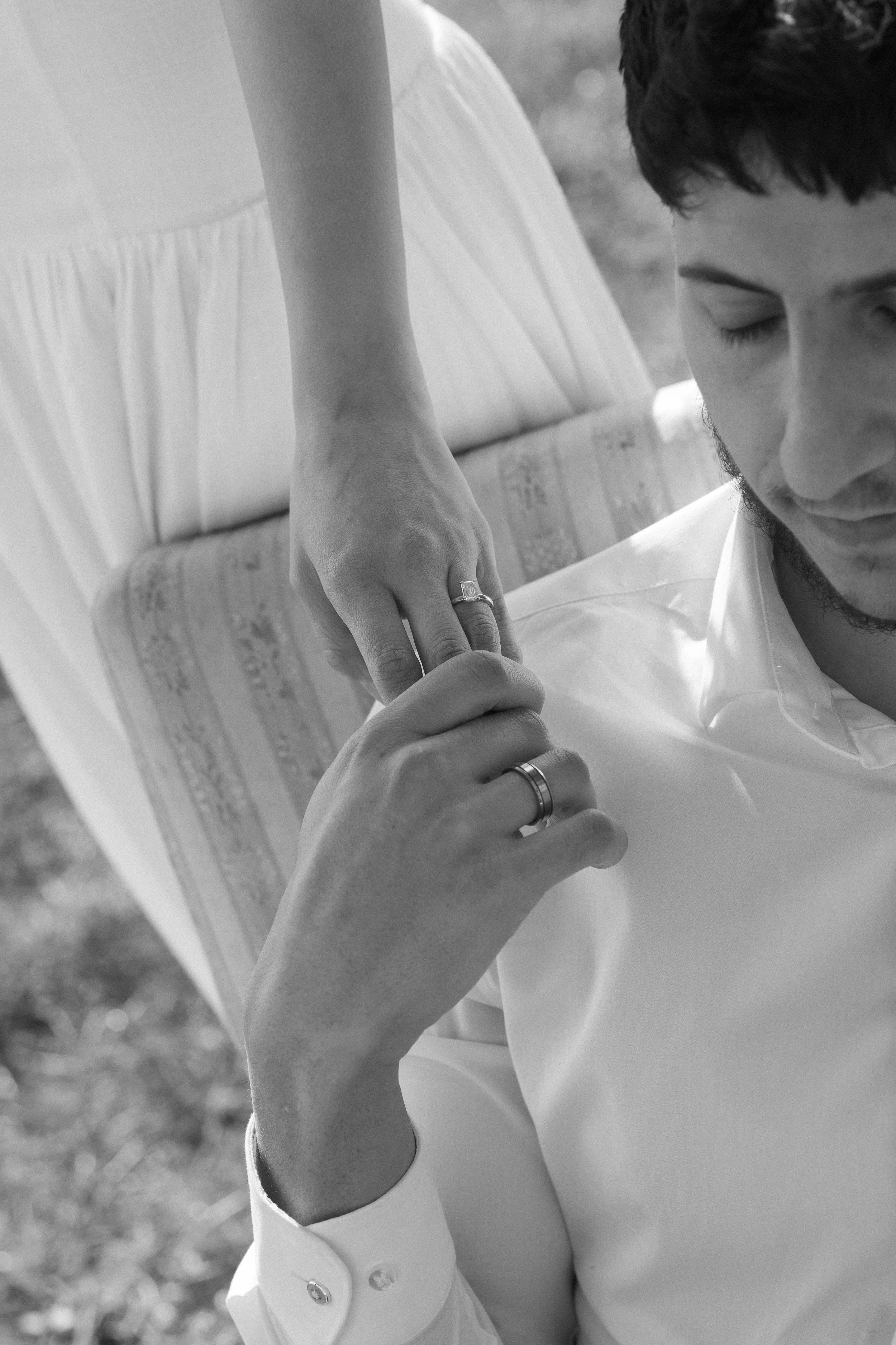 A black and white photo of a man with a beard and dark hair, wearing a white shirt, with a woman standing behind him. The woman's hand rests on the man's shoulder, showing a ring on her finger, and is dressed in a light-colored dress. The woman is out of focus, and the man has his eyes closed.