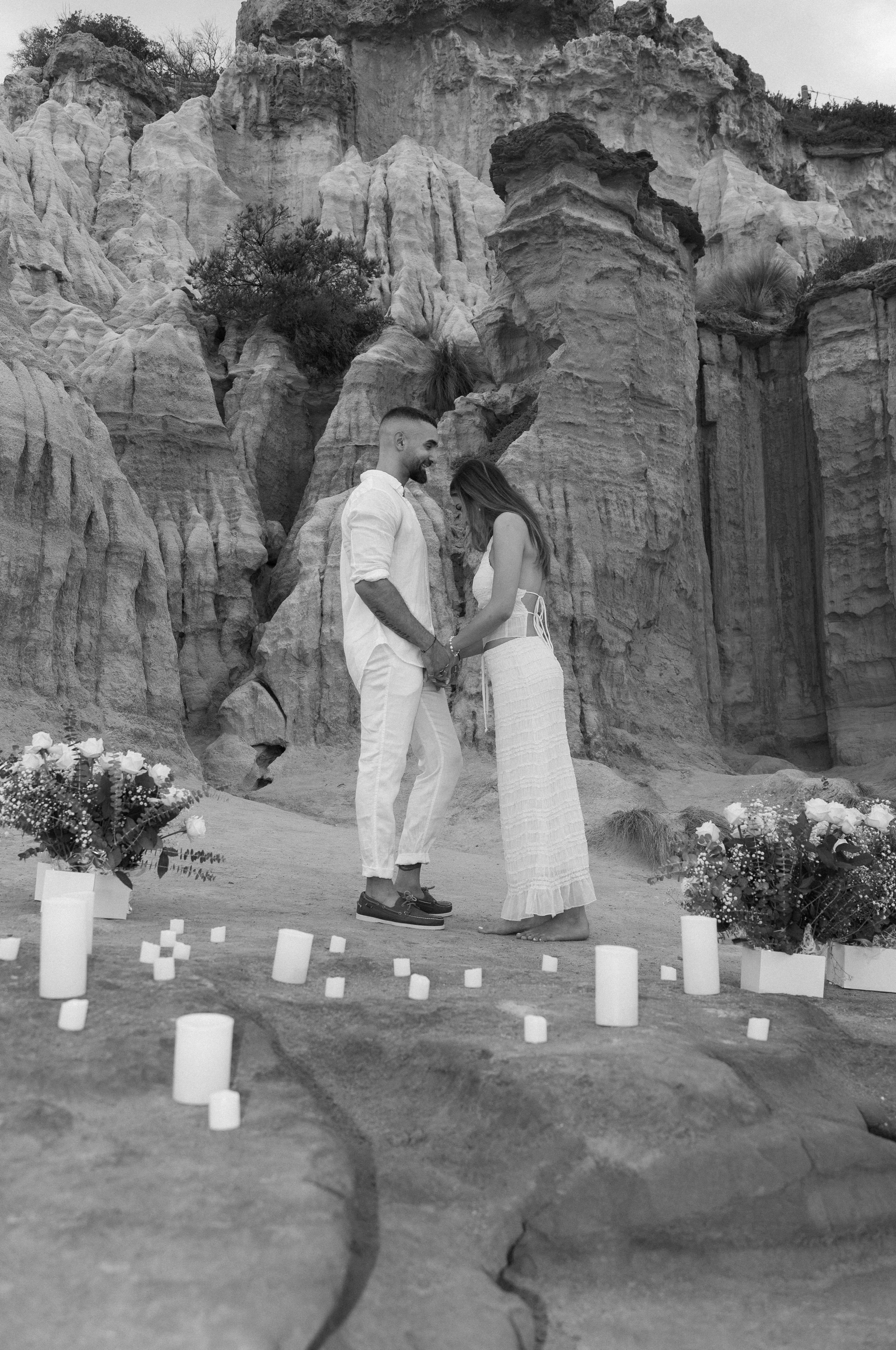 A couple holding hands and smiling at each other during a wedding ceremony outdoors, with a rocky canyon background and lit candles and flower arrangements on the ground.