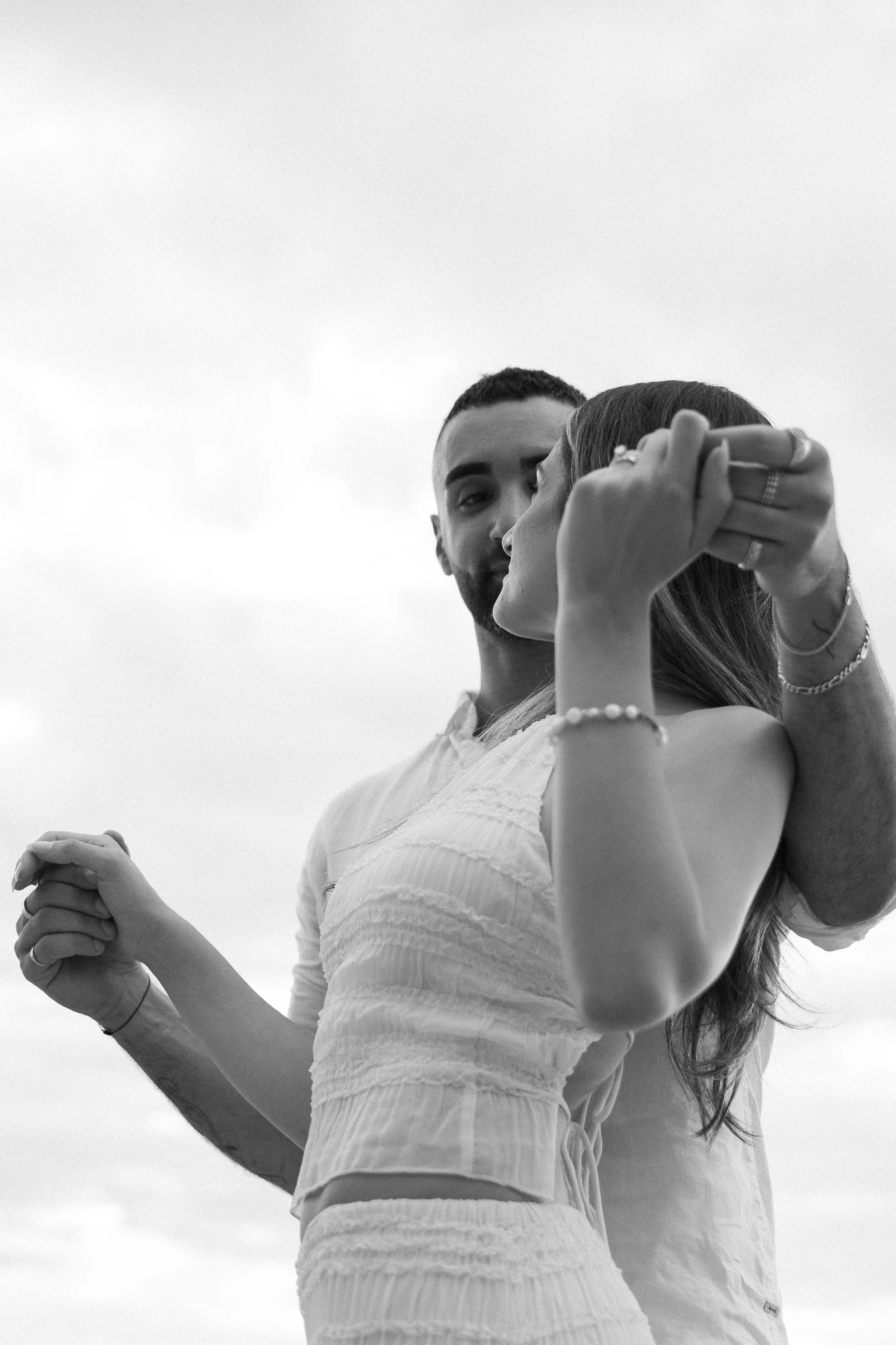 A black and white photo of a couple kissing, with the man standing behind the woman, holding her head gently, against a cloudy sky background.
