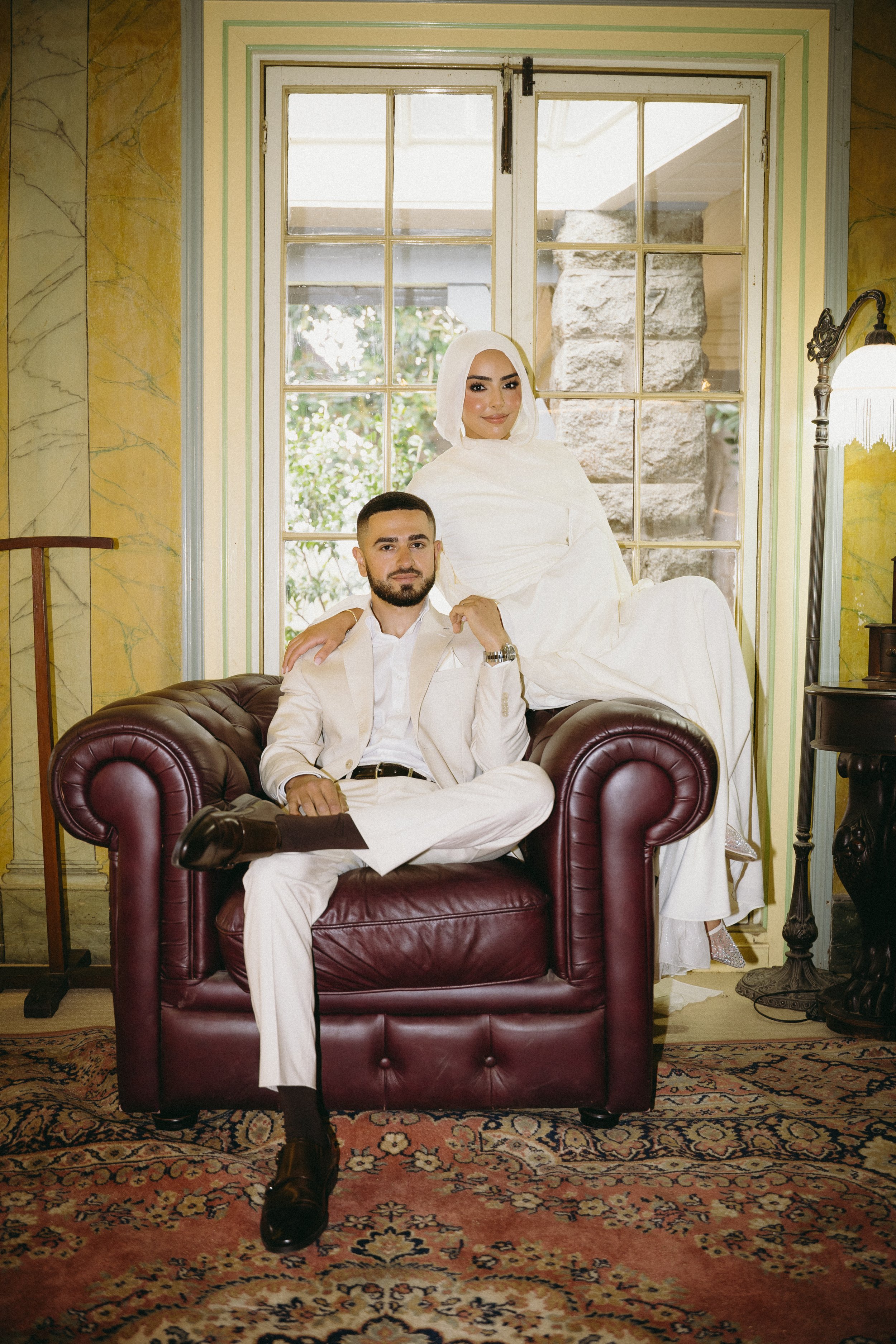 A man dressed in a white suit and a woman dressed in a white dress with a headscarf pose together in a vintage-style room, sitting on a leather sofa with a large window behind them.