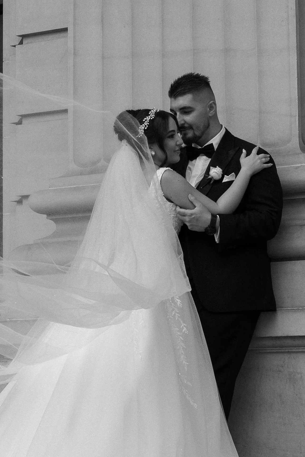 A black and white photo of a bride and groom embracing each other, with the bride wearing a wedding dress and veil, and the groom in a tuxedo, standing against a stone wall.