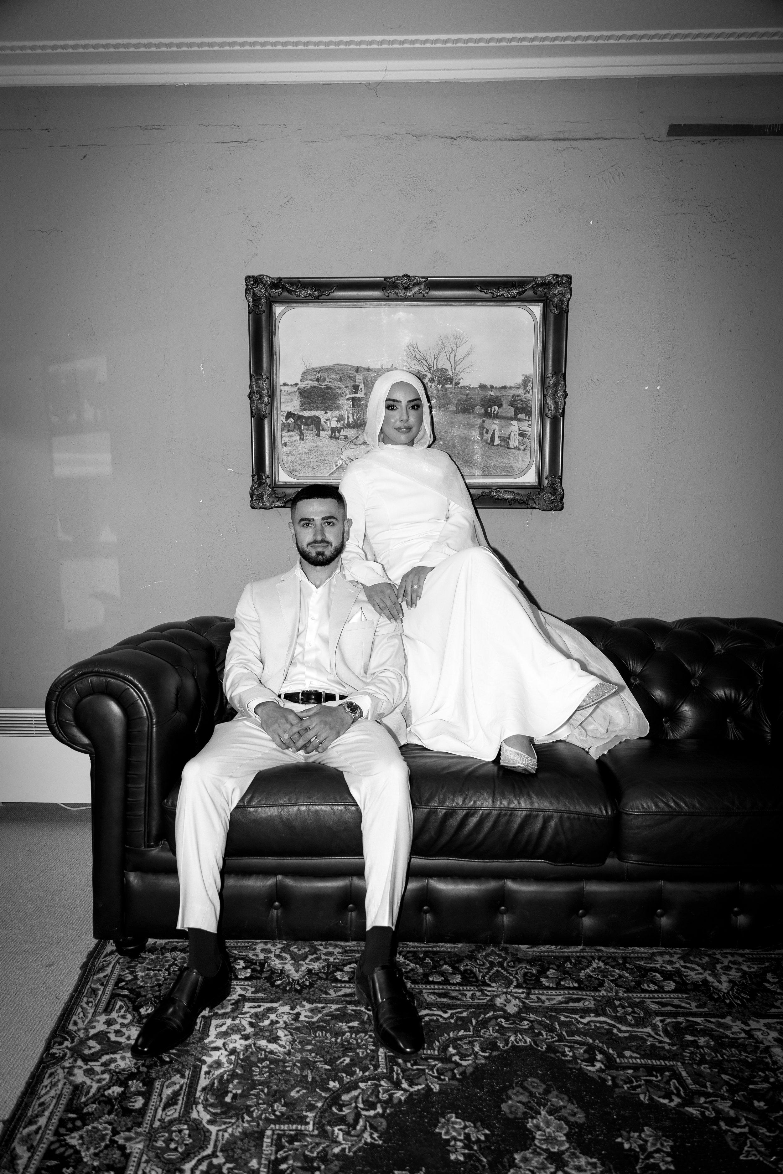 A black and white photo of a man and woman dressed in formal attire sitting on and standing behind a leather sofa in a room with a framed painting of a rural scene behind them.