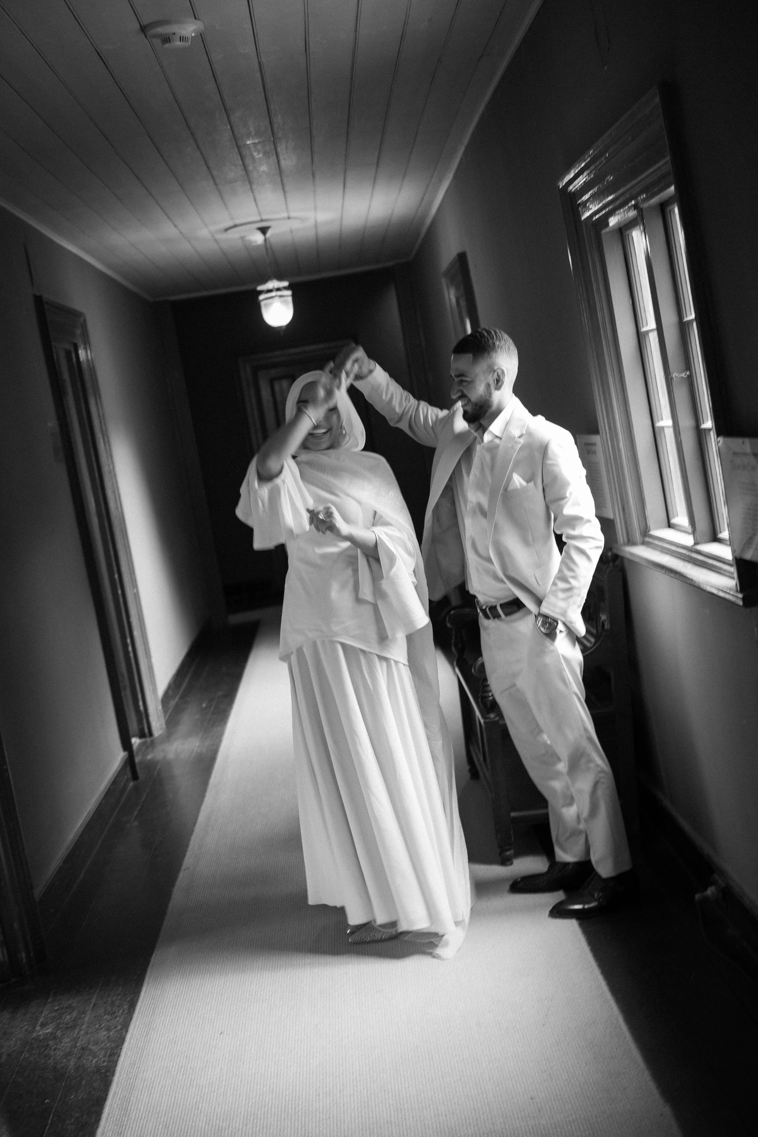 A black and white photo of a woman in a long dress with a shawl dancing with a man in a suit in a hallway with windows.
