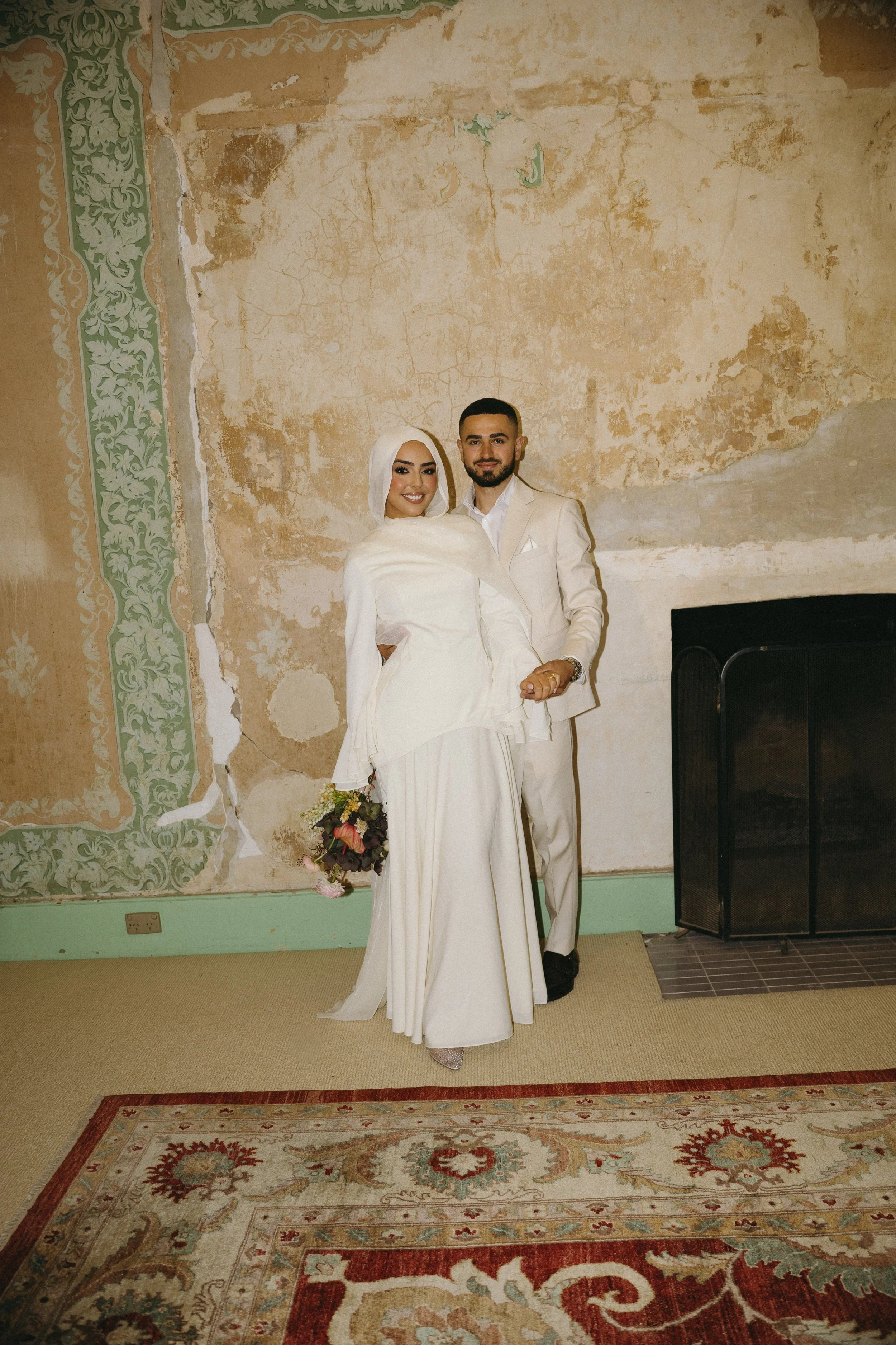 A bride and groom in white wedding attire, standing together indoors in front of a worn, textured wall and a fireplace, with a floral carpet in the foreground.