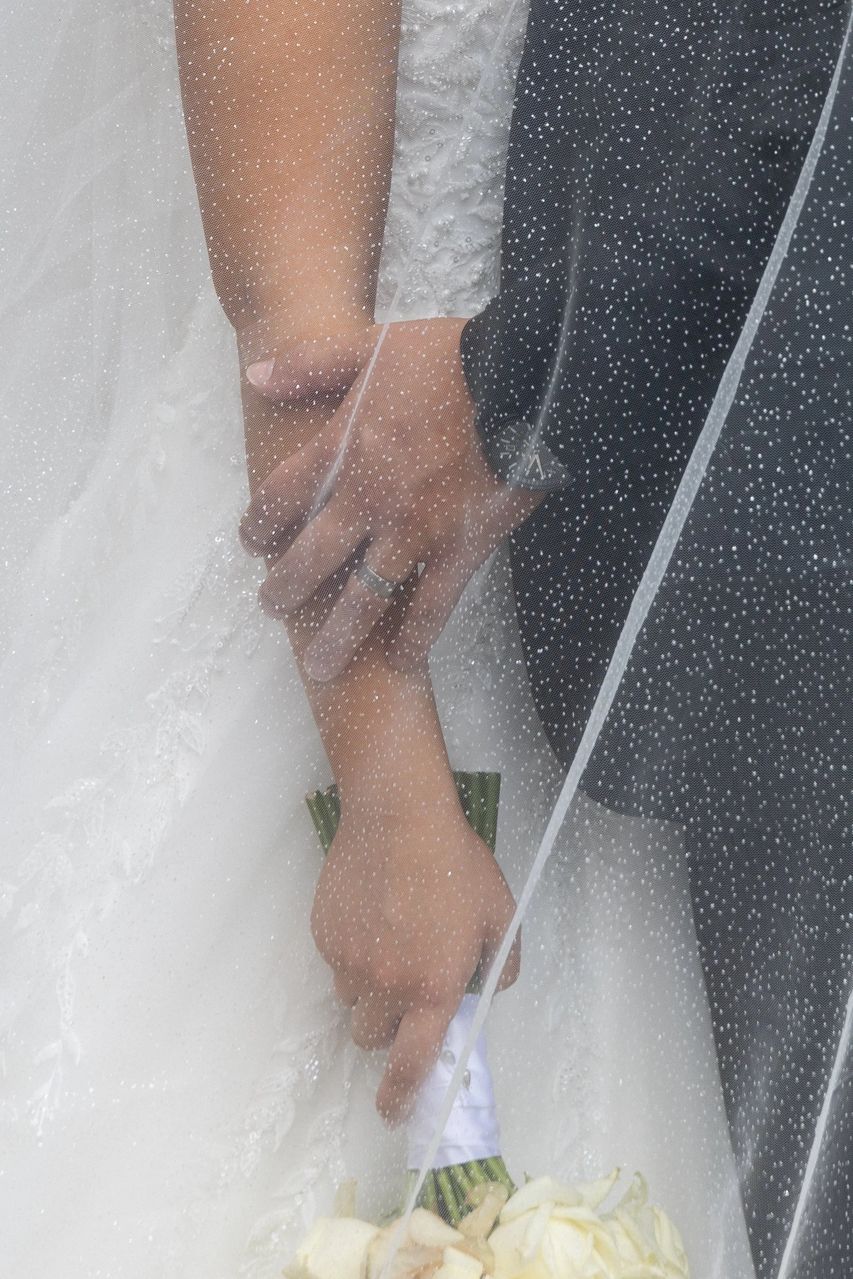 Close-up of a bride and groom holding hands during their wedding ceremony, with the bride in a white wedding dress and the groom in a dark suit, photographed through a rain-covered glass window.