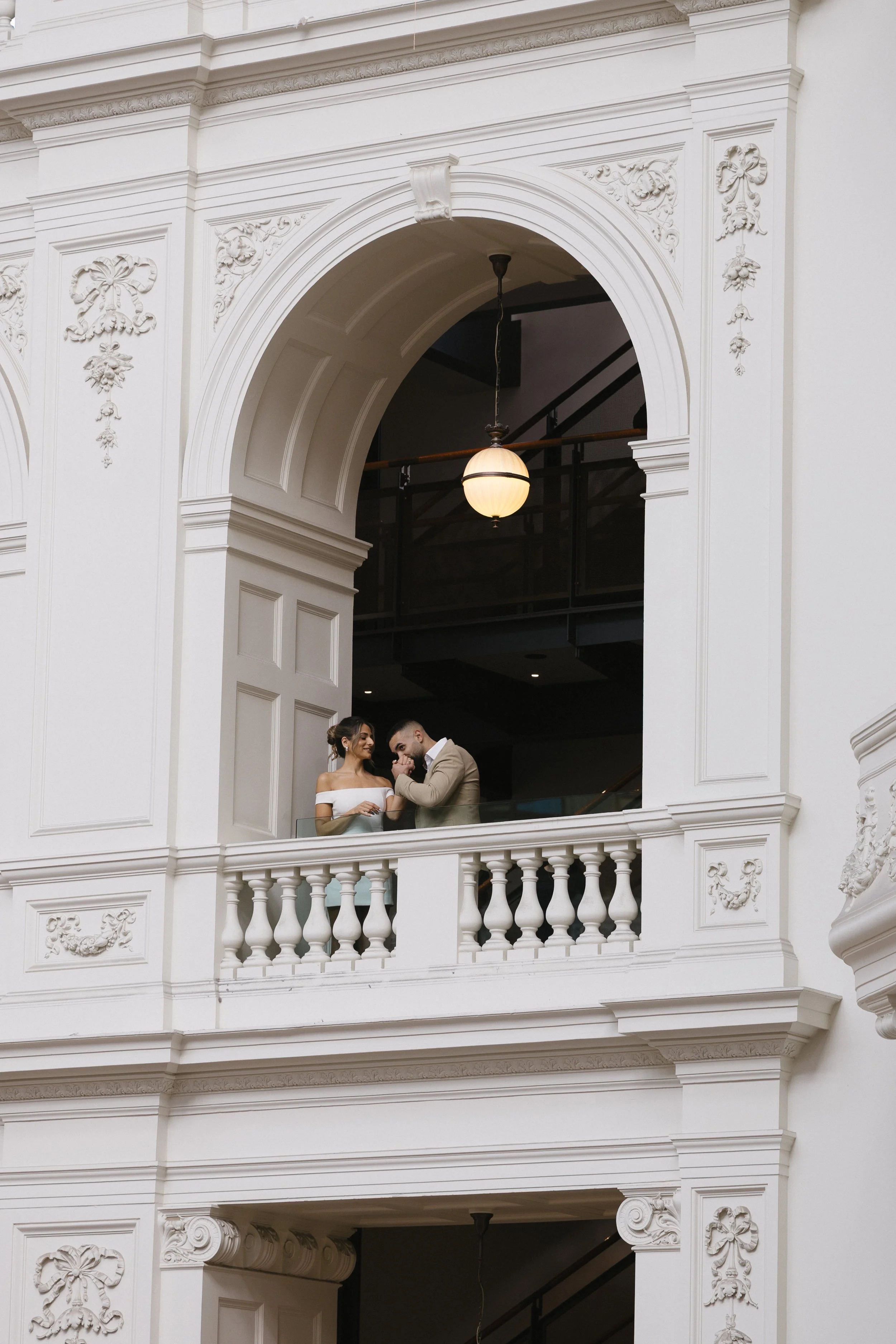 A a woman and man stand on a balcony inside a building with ornate white decor. The woman is wearing a white off-shoulder dress, and the man is dressed in a light-colored suit. They are holding hands and appear to be whispering or sharing a secret.