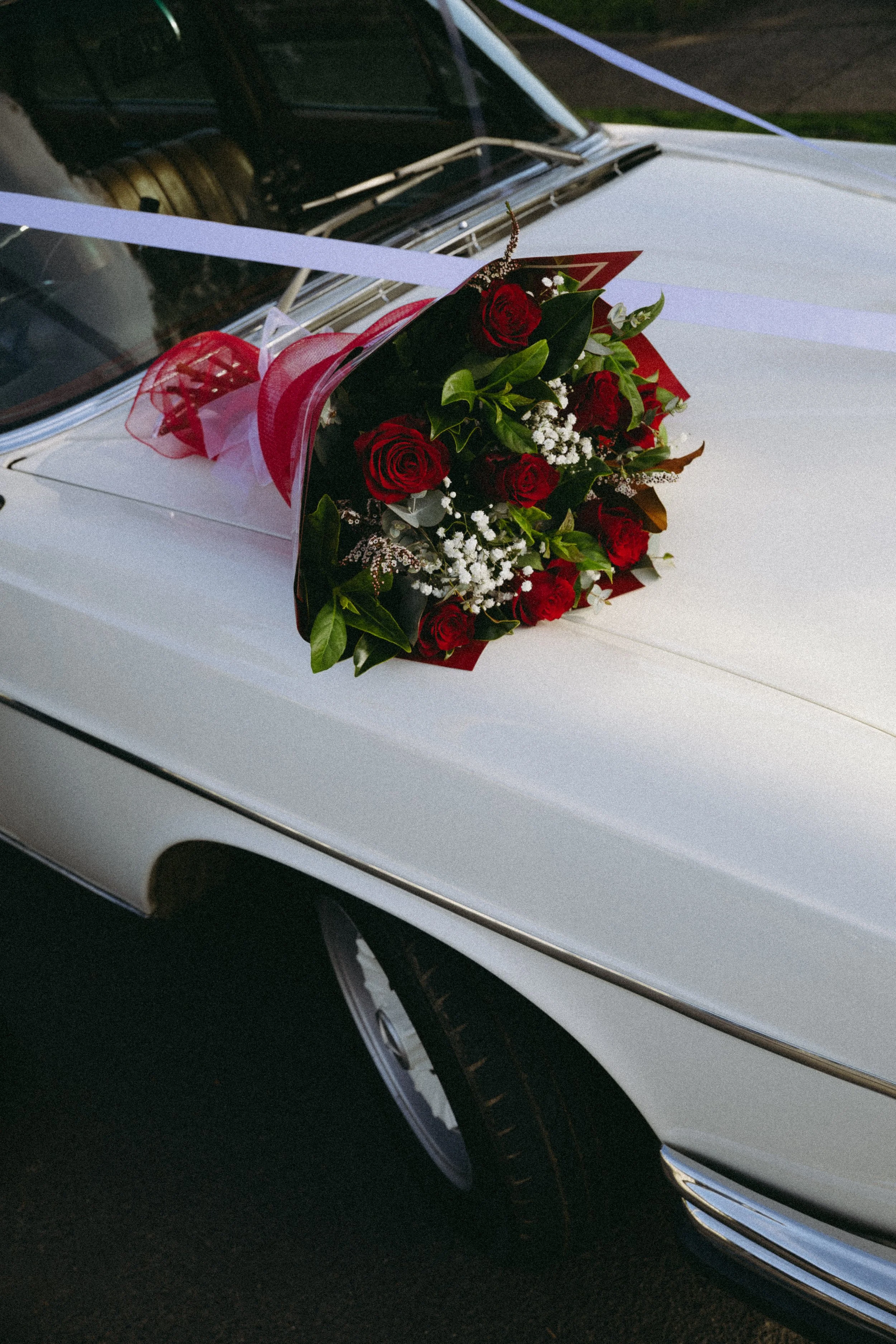 White vintage car with a bouquet of red roses and white flowers on the hood, decorated with white ribbons.
