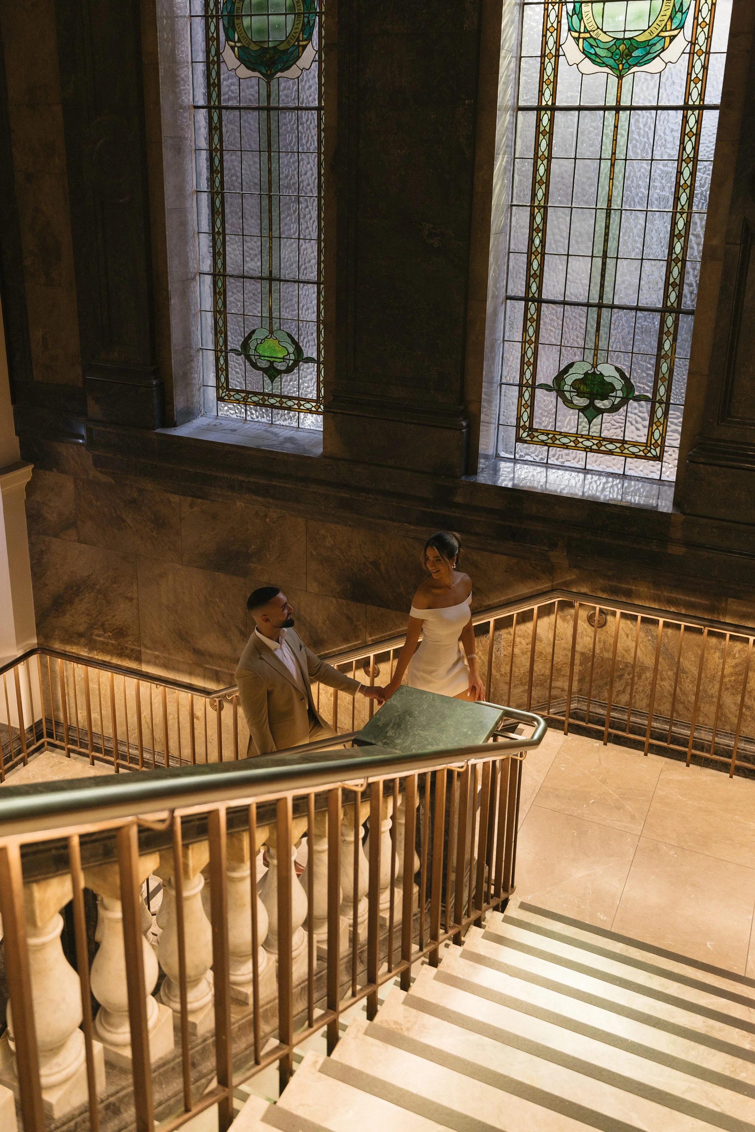 A man and woman holding hands on a staircase inside a building with stained glass windows, marble walls, and a tiled floor.