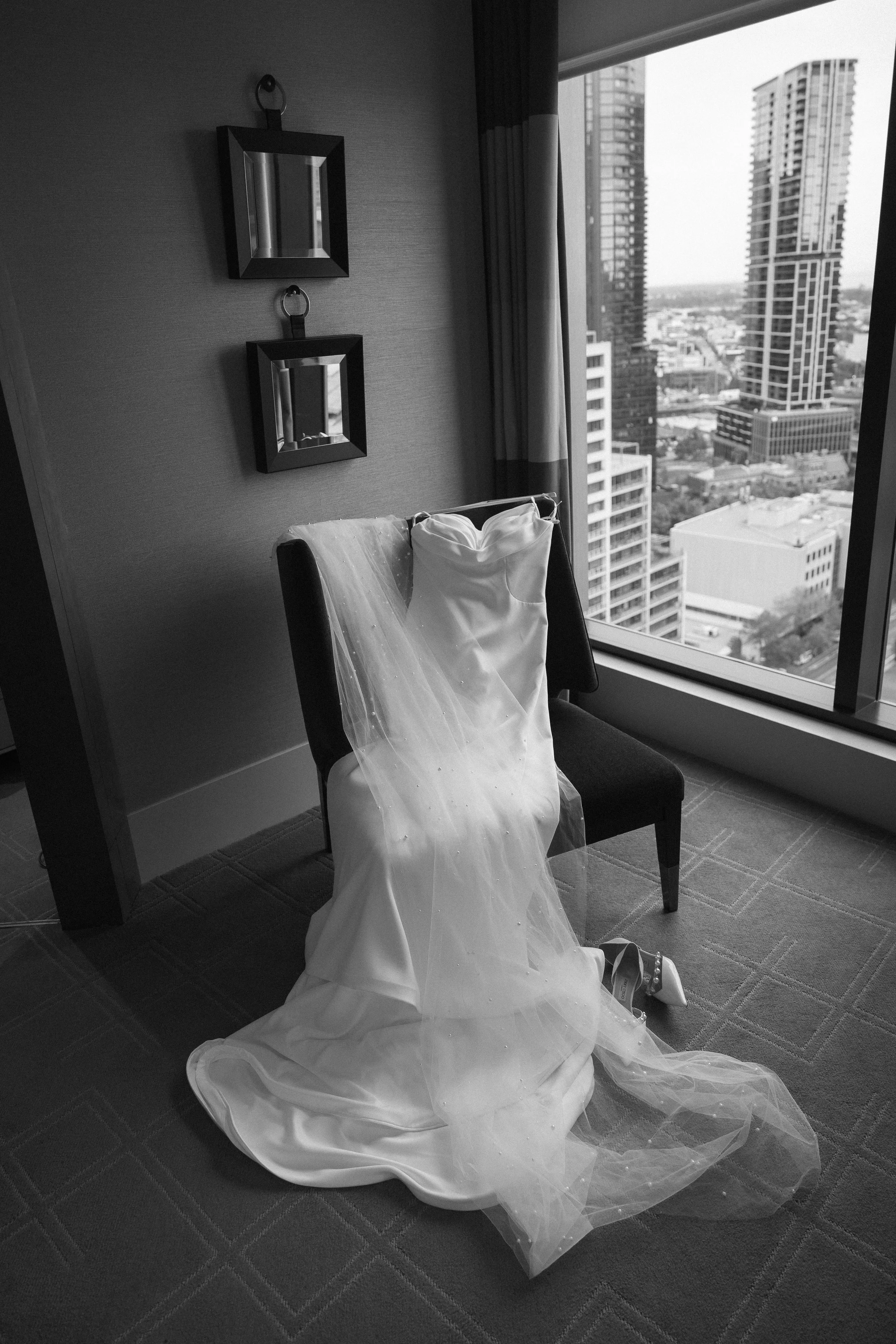 Black and white photo of a wedding dress hanging on a chair near a large window with a city skyline viewed outside.