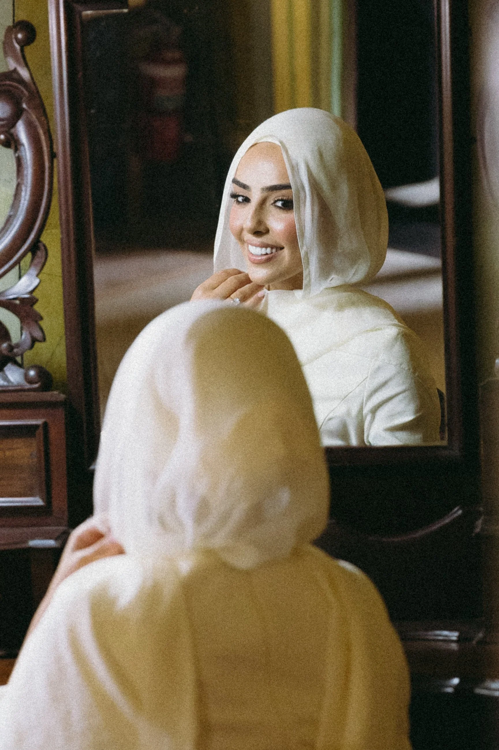 A woman in a light-colored hooded outfit looking at herself in a mirror, smiling and touching her chin.