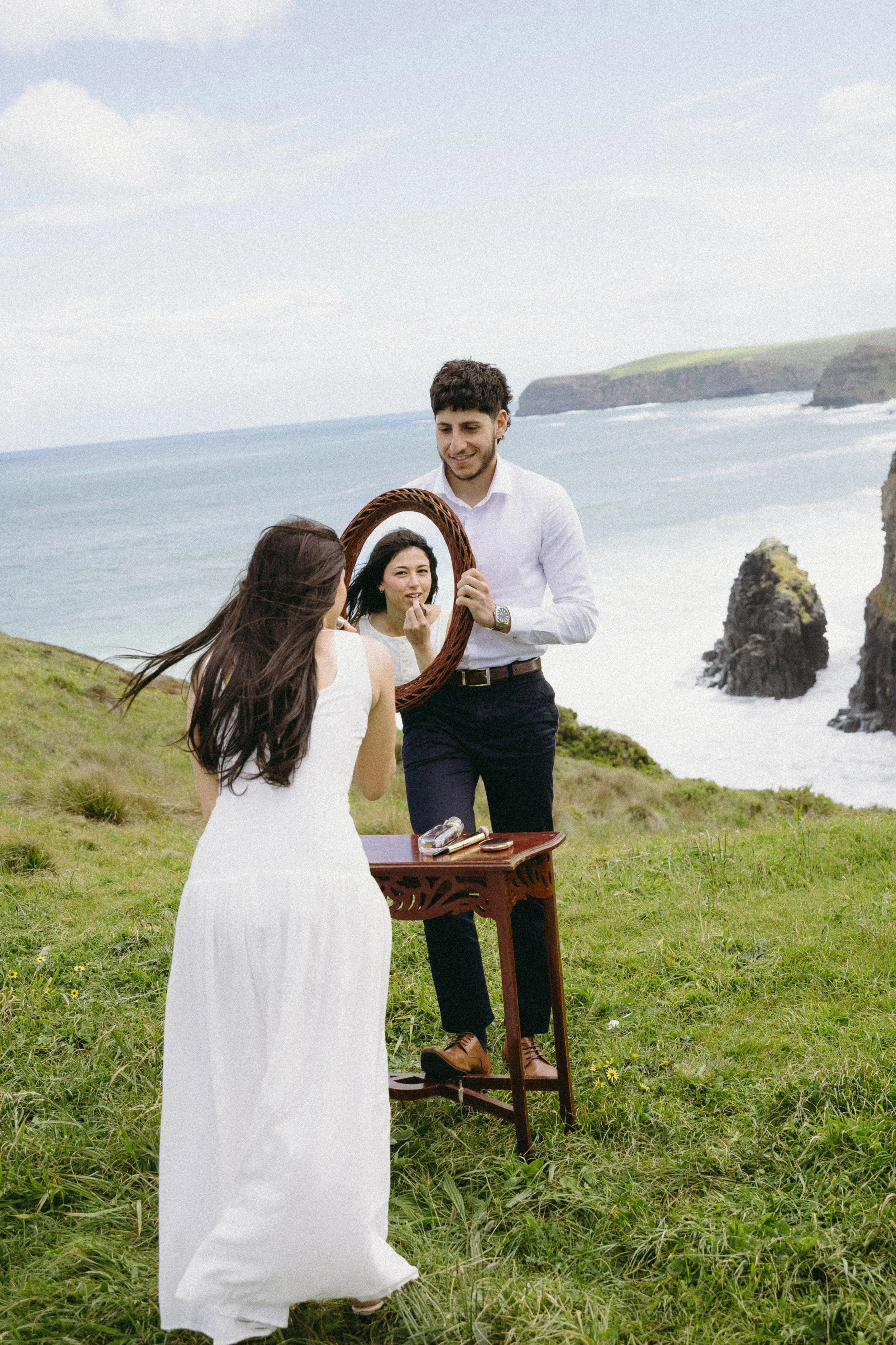 A woman in a white dress stands outdoors on grass, looking into a mirror held by a man in a white shirt. They are on a grassy hill near the coast with rocky cliffs and the ocean in the background.
