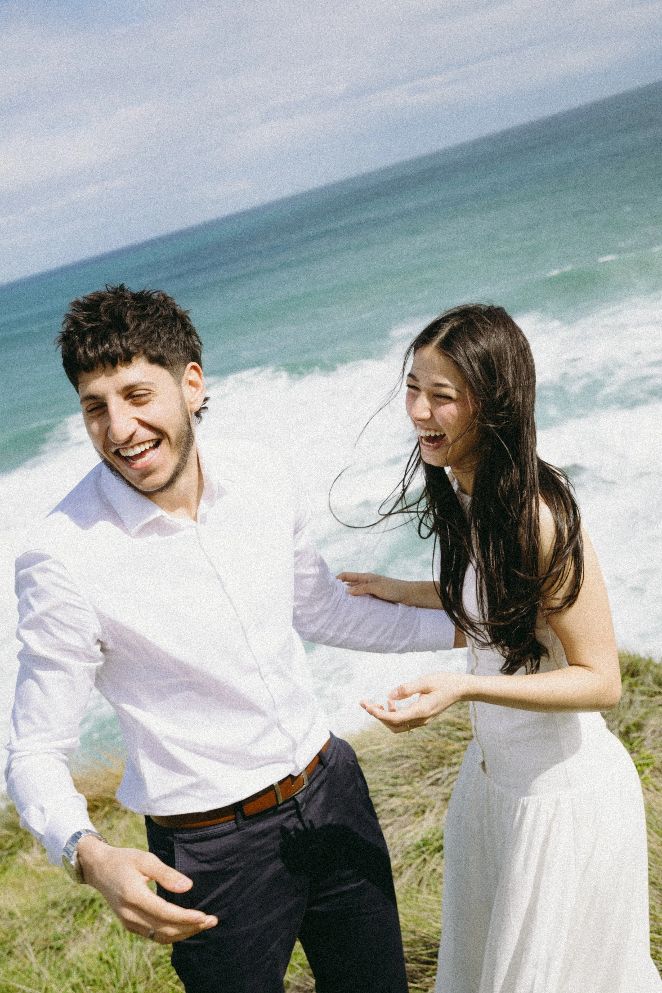 A young man and woman laughing and enjoying time together on a grassy area near the ocean, with waves and sky in the background.