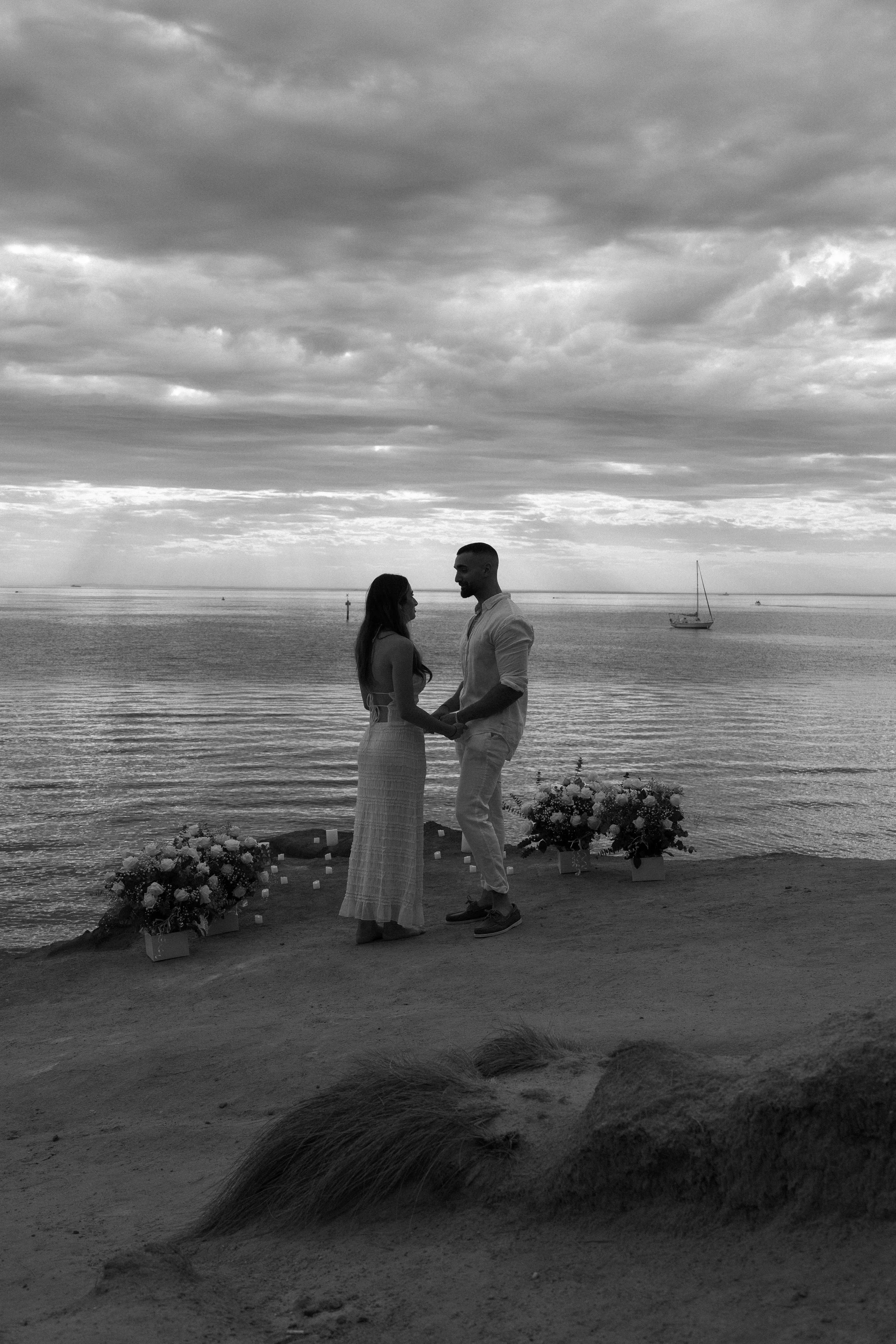 A couple standing on a beach holding hands, facing each other during sunset or sunrise, with a sailboat in the water in the background and flowers arranged on the sand around them.