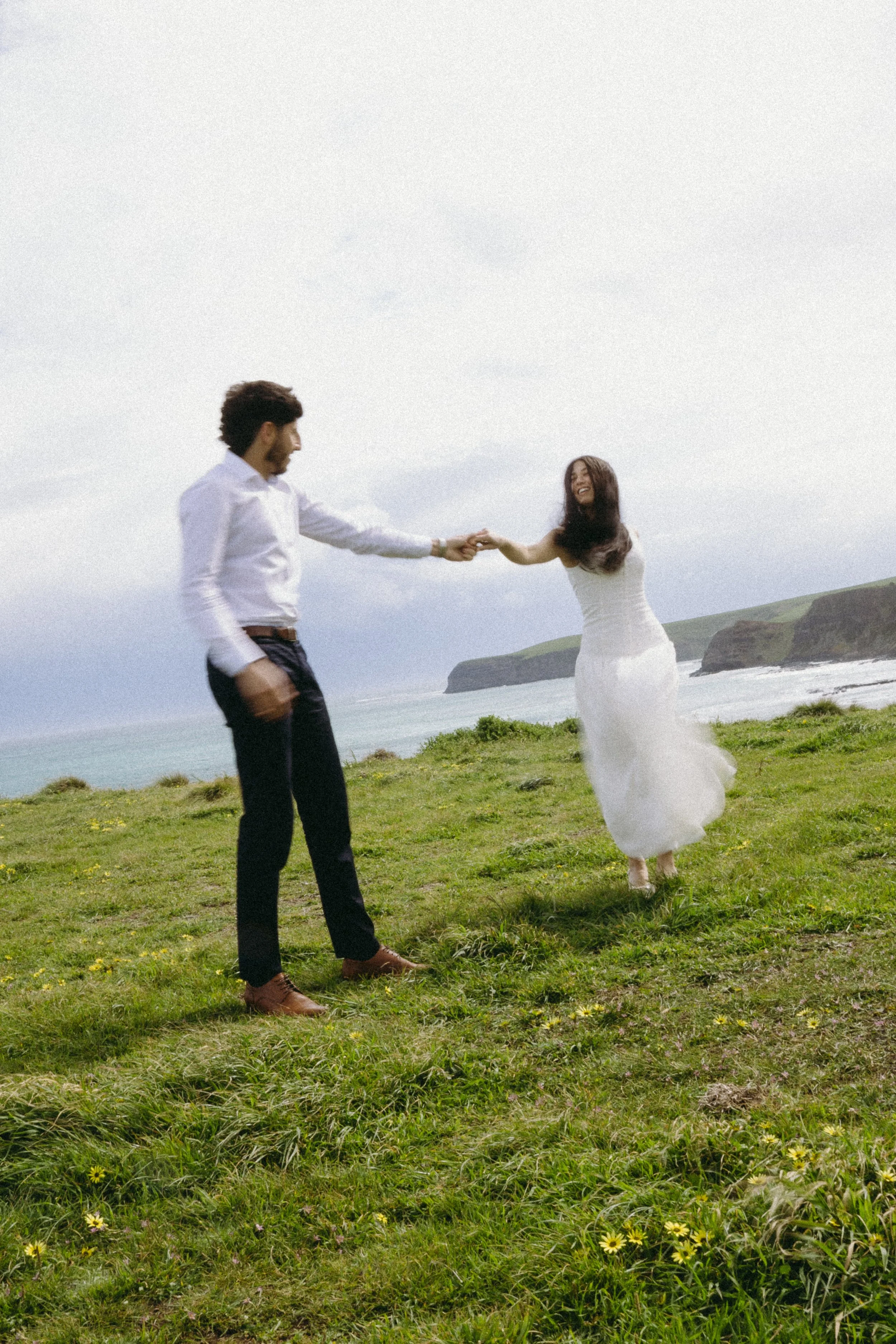 A couple holding hands and laughing on a grassy cliffside overlooking the ocean and rocky cliffs.