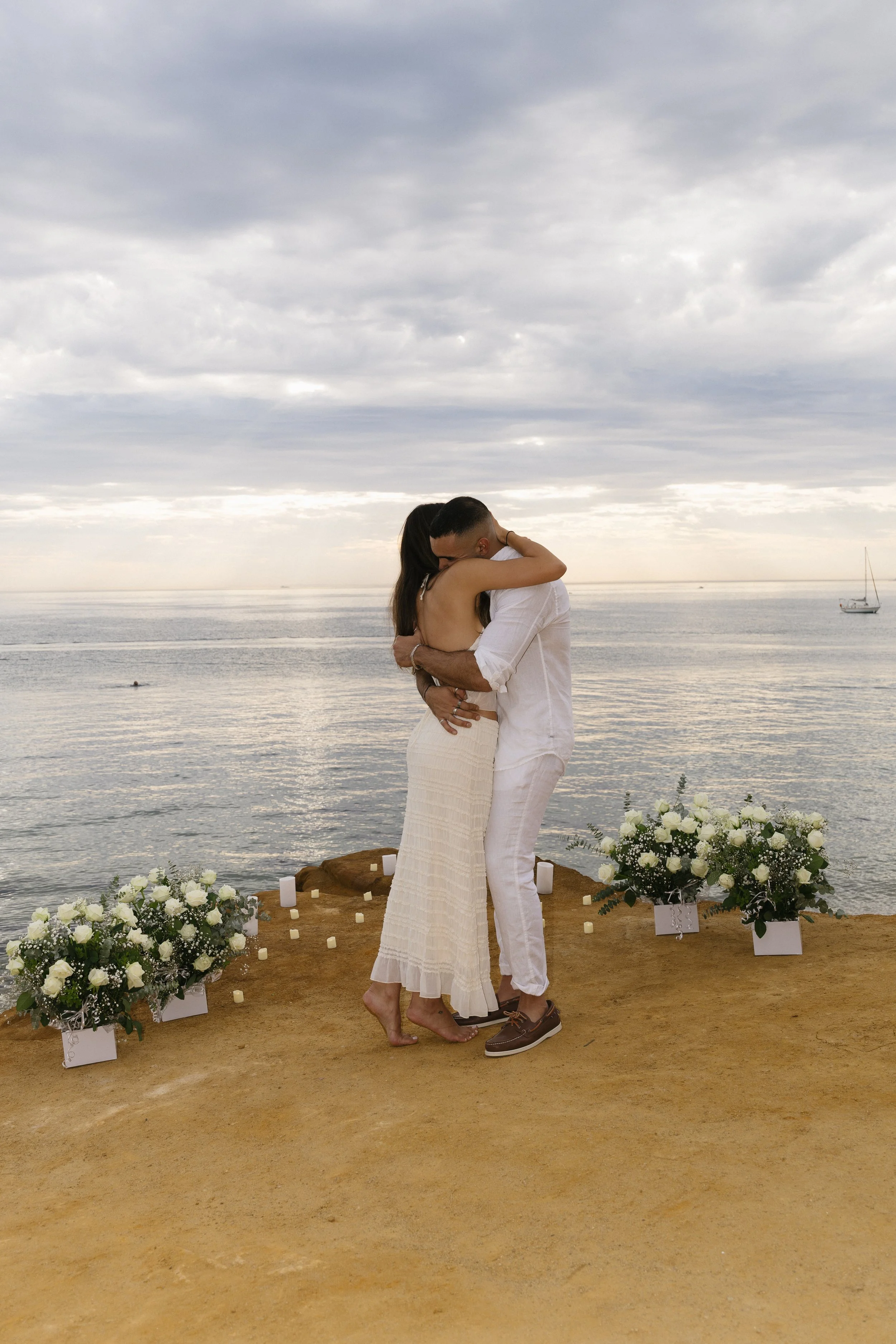 A couple hugging on a beach during sunset with floral arrangements and candles around them, possibly exchanging vows.