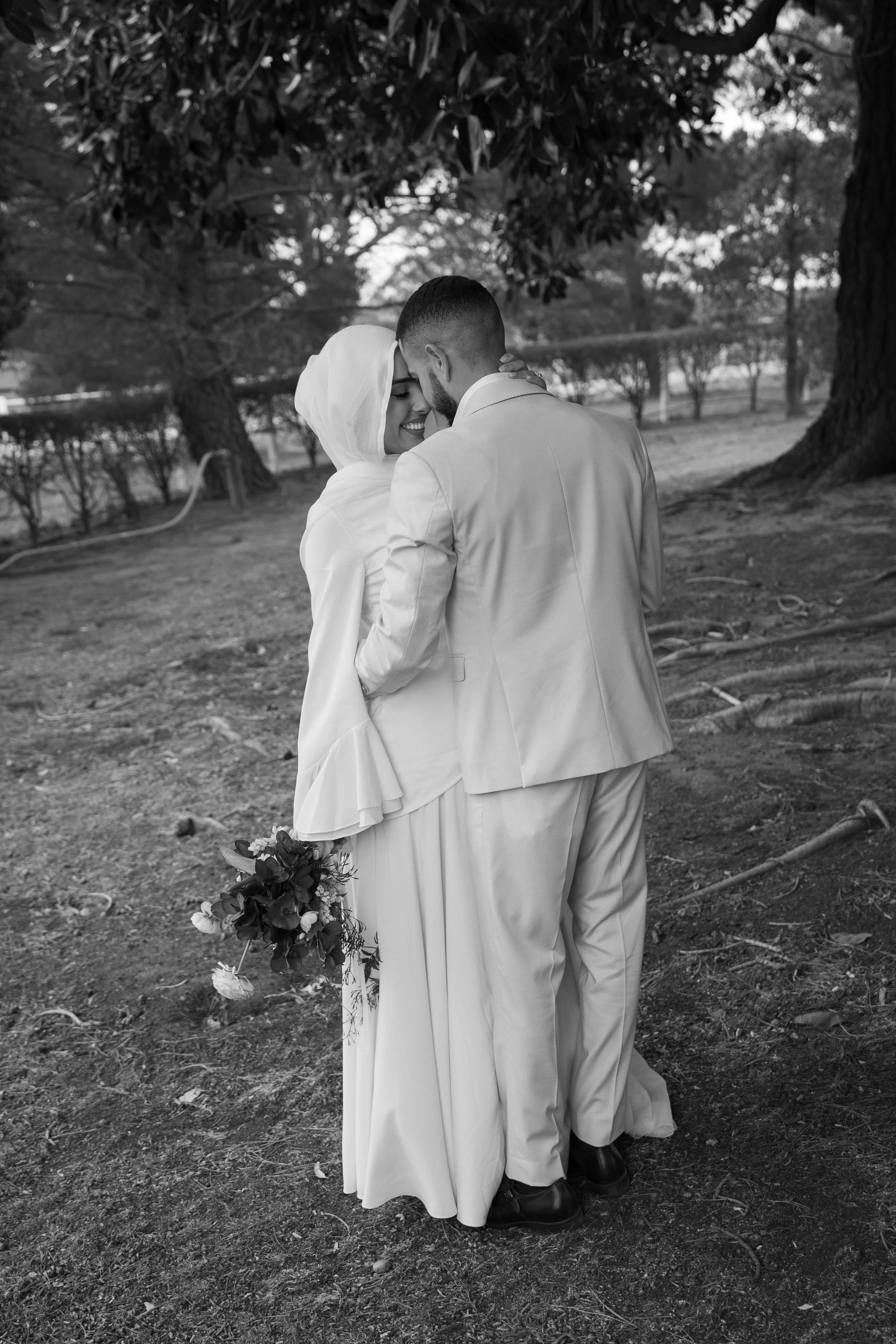 A black and white photo of a couple dressed in wedding attire embracing outdoors under a large tree, with trees and open space in the background.