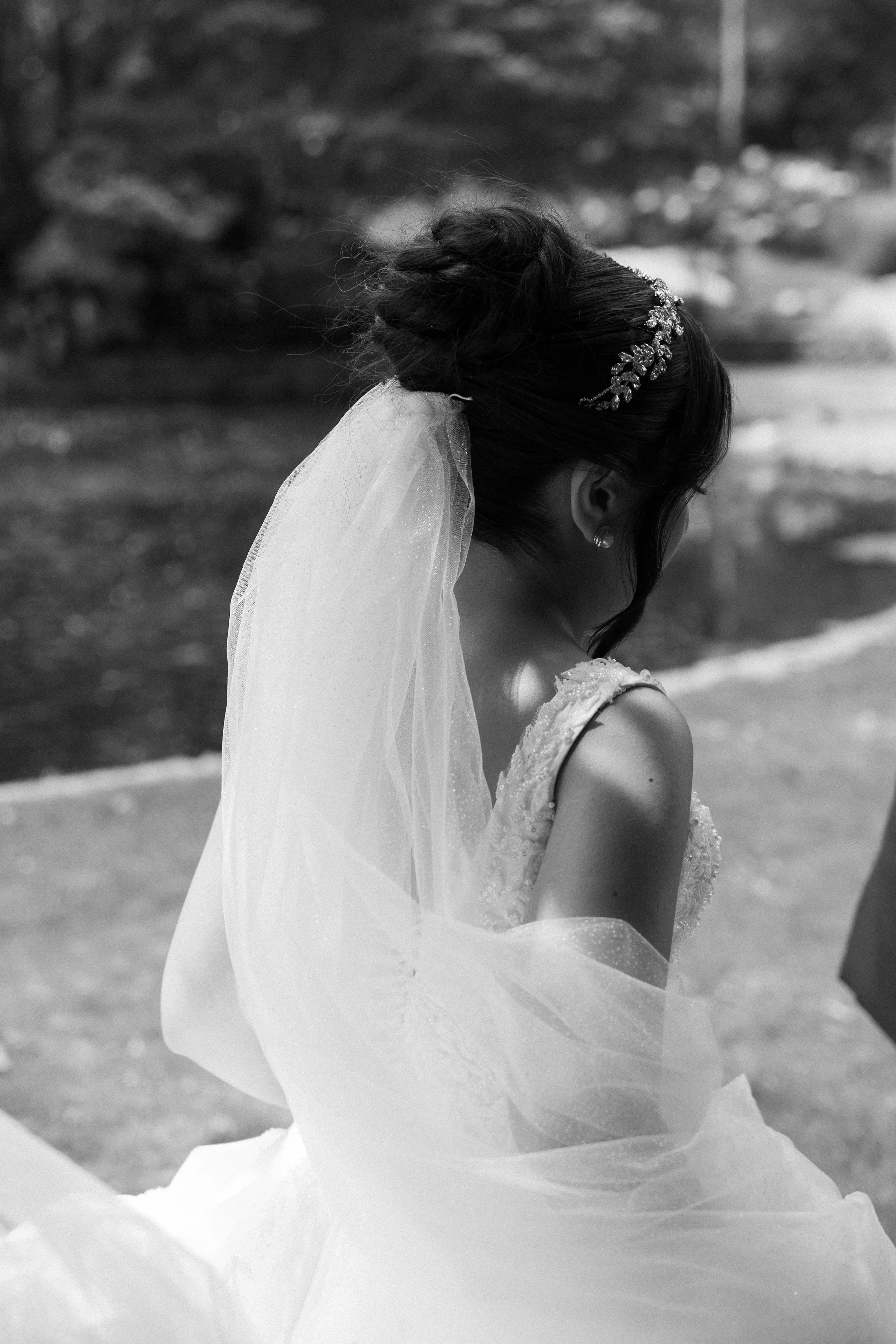 A bride in a wedding dress with a veil and hair styled in an updo with a decorative hairpiece, standing outdoors.