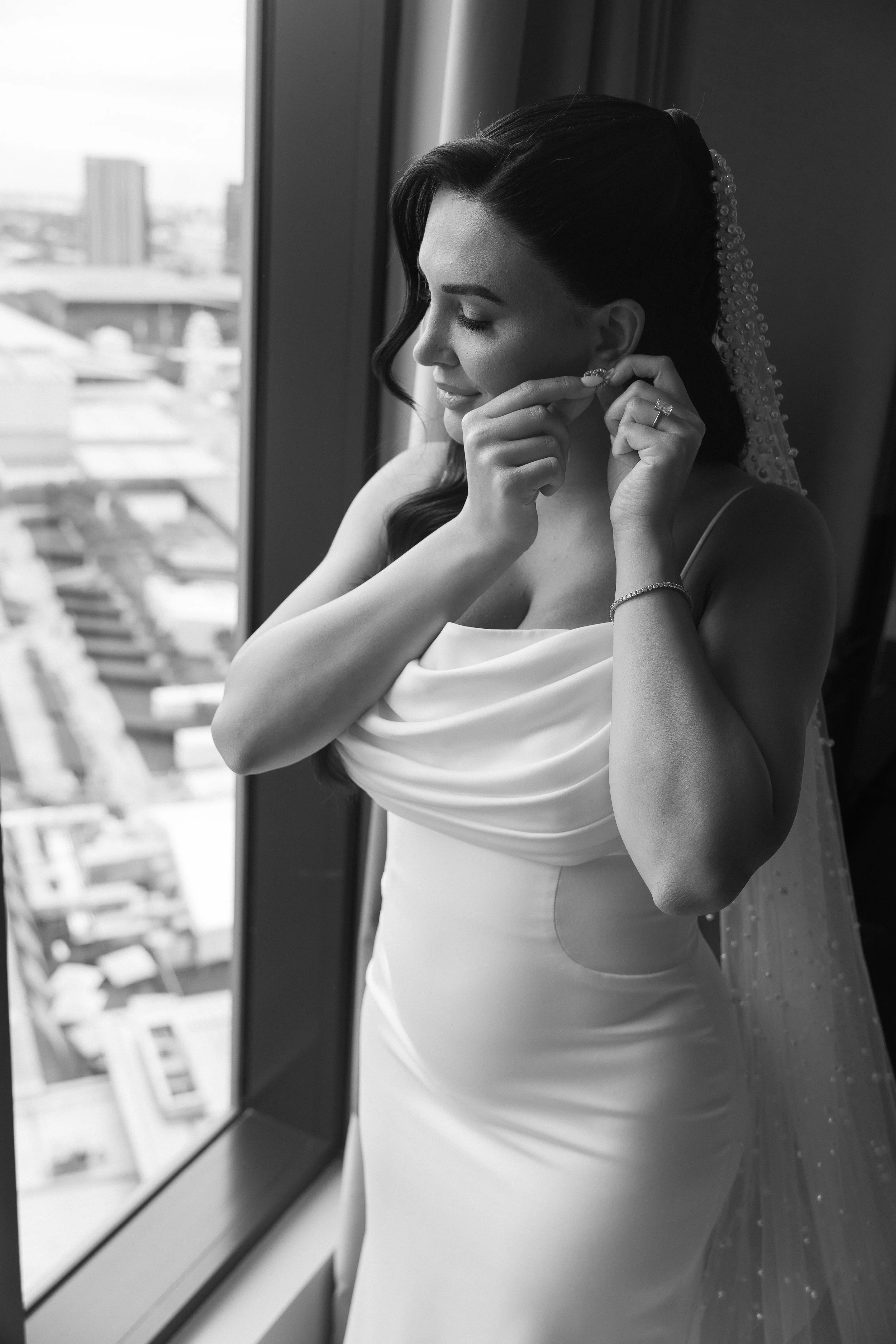 A bride standing by a window, adjusting her earring, wearing a wedding dress with a fitted bodice and draped neckline, and a veil with pearl embellishments.