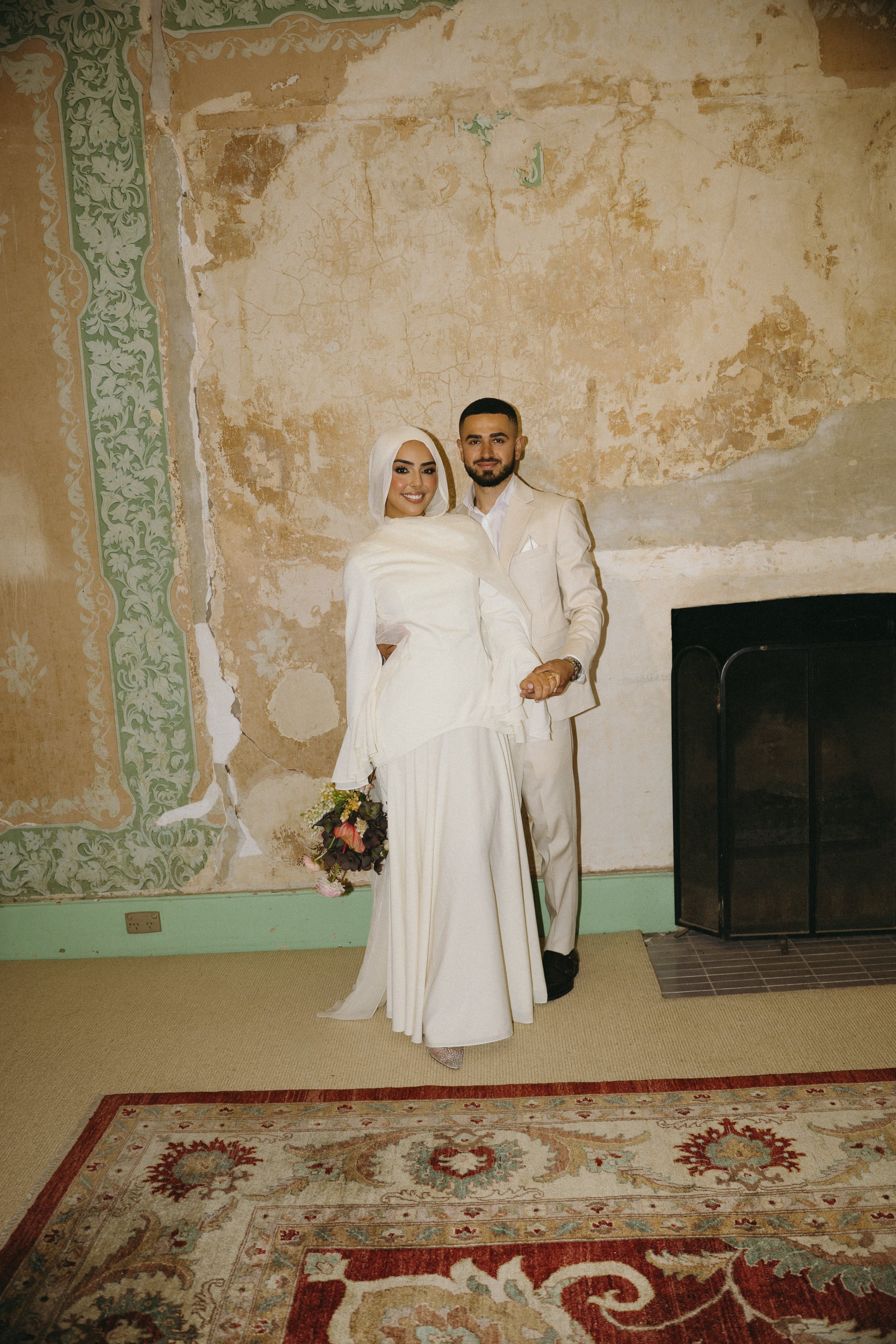 A bride and groom stand together indoors, smiling, with the bride holding a bouquet. The wall behind them is old and peeling with decorative green and beige paint, and there is a fireplace to the right.