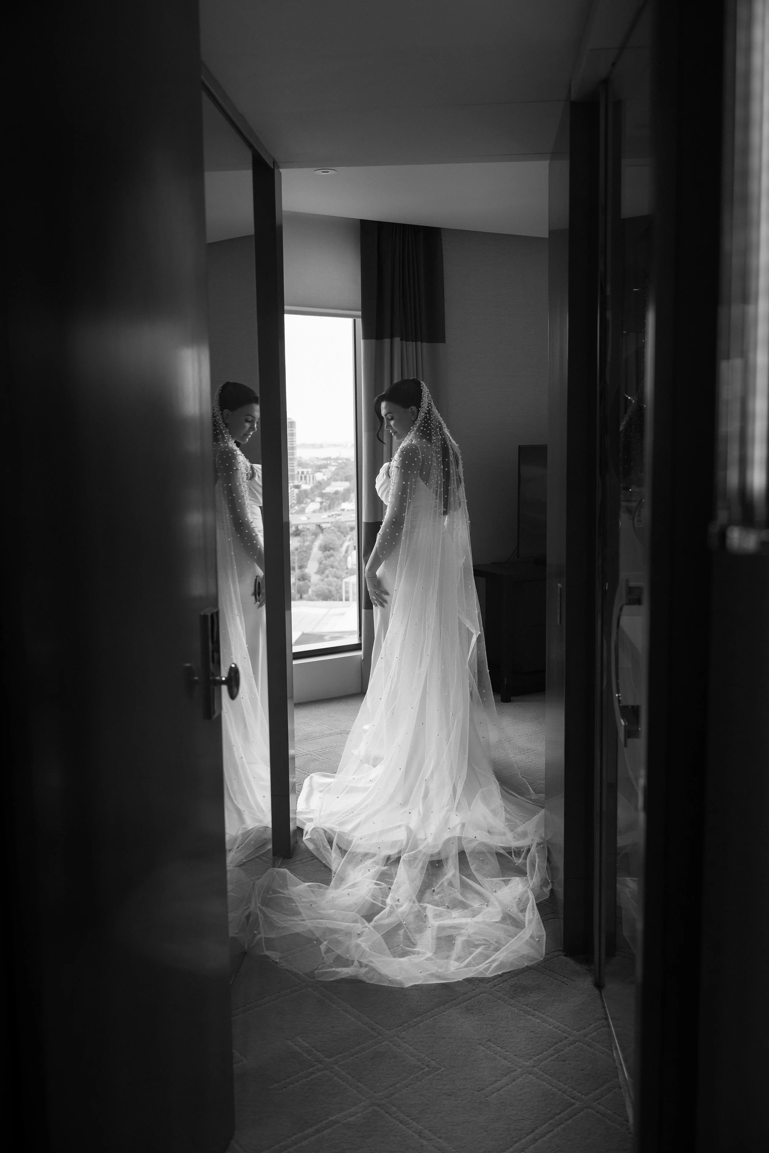 A bride in a wedding dress with a veil stands in a hotel room, looking down, reflected in a mirror, with a window and cityscape in the background.