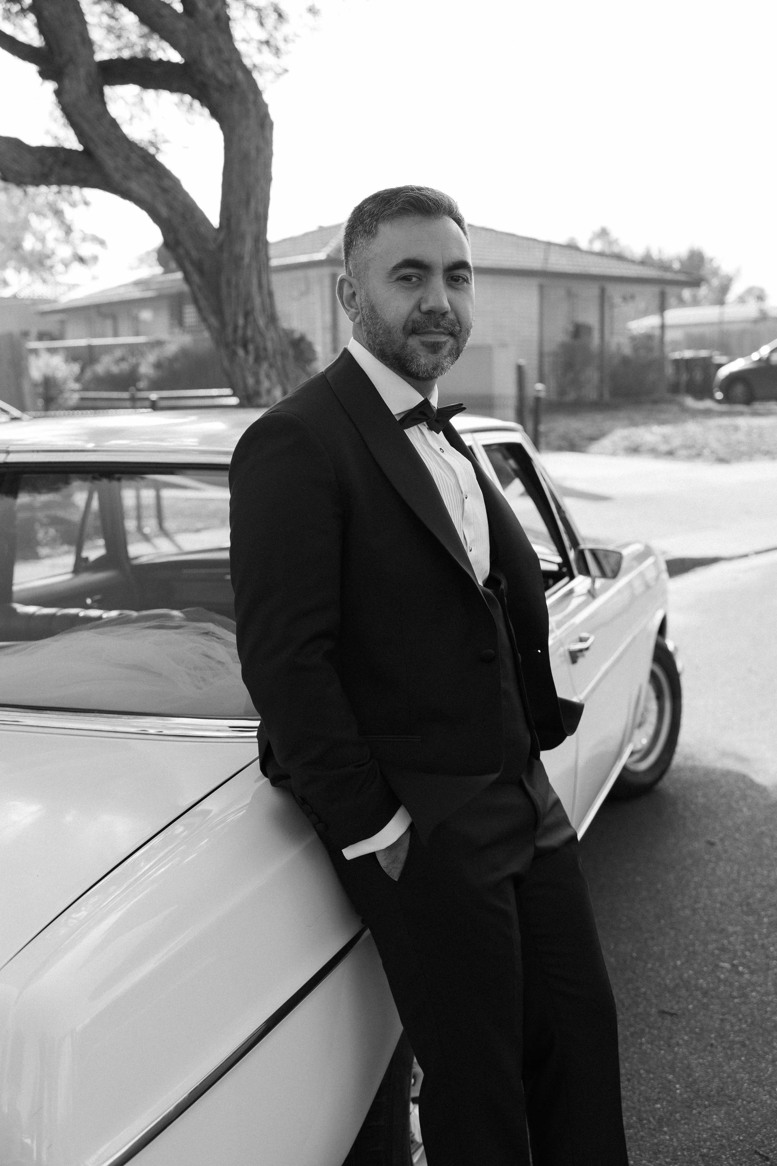 A man in a tuxedo leaning against a vintage car outdoors in a residential neighborhood.