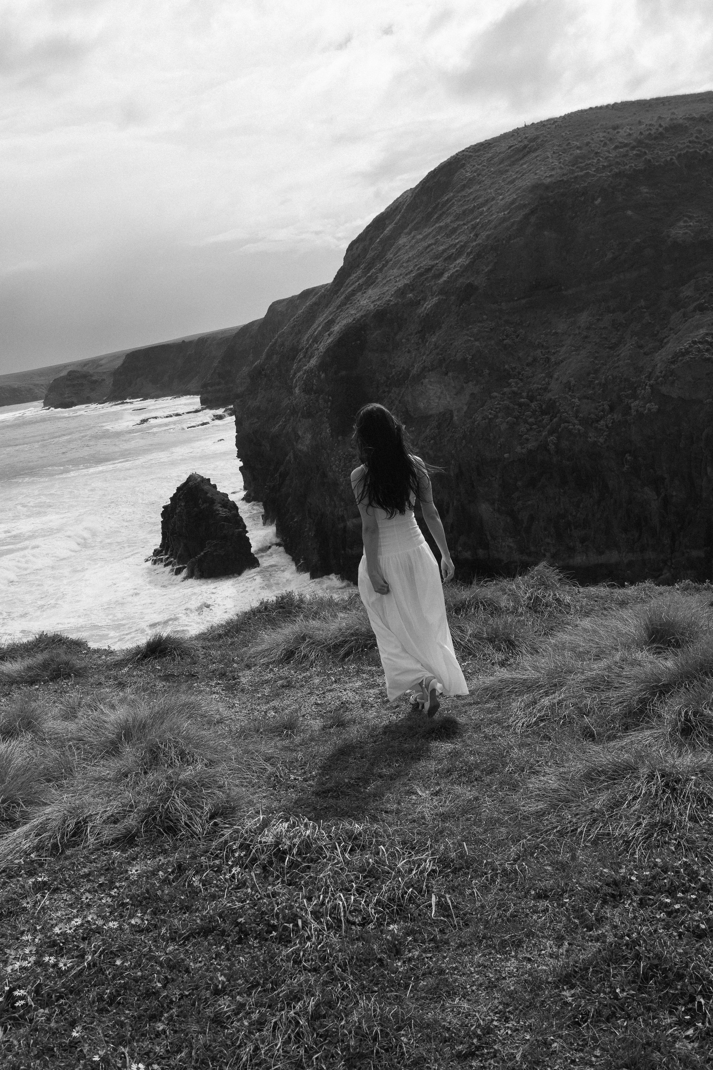 A woman in a white dress walking along a grassy cliff by the ocean, with large rocks and cliffs in the background, in black and white.