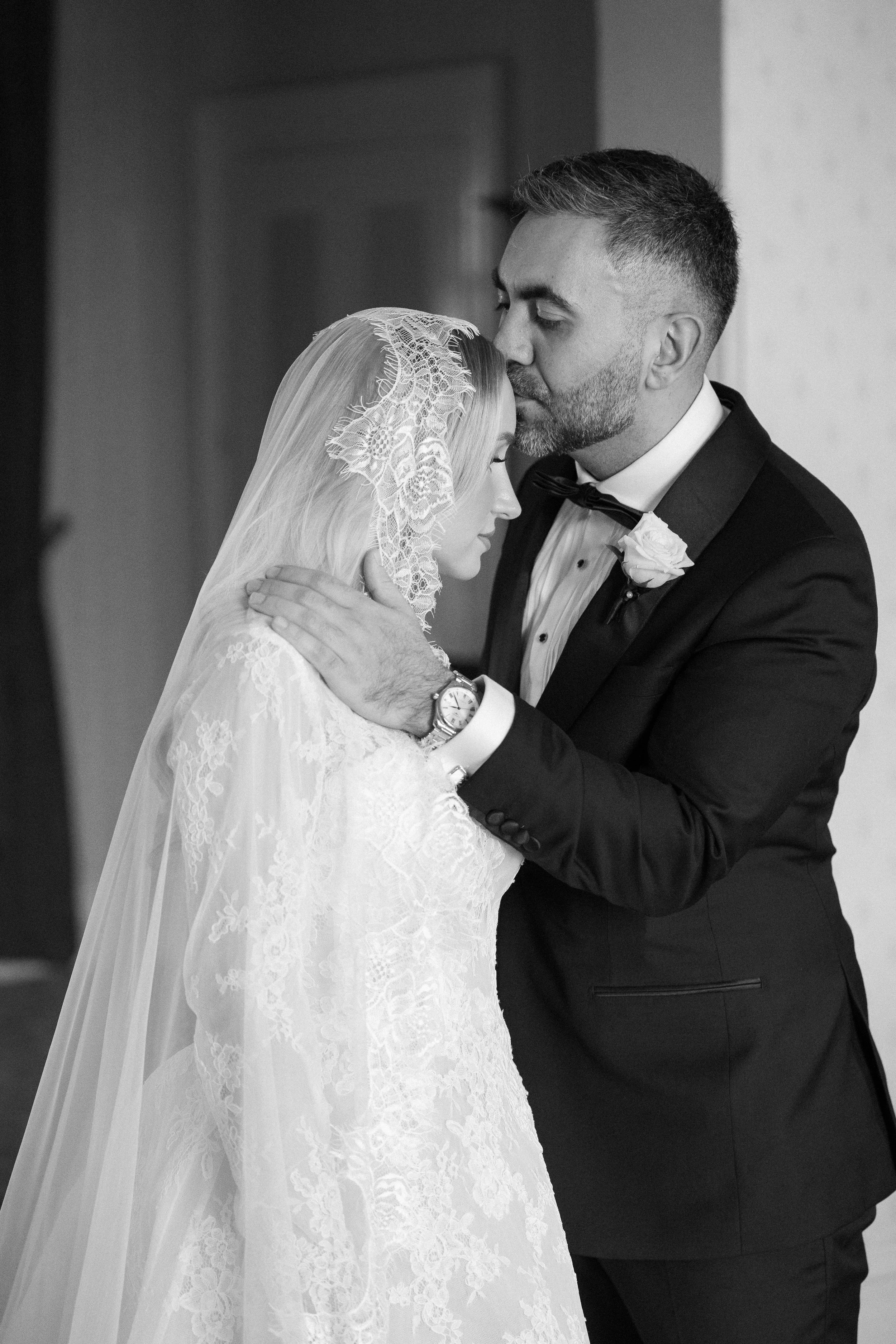 A man and woman sharing an intimate moment on their wedding day, with eyes closed and foreheads touching, dressed in wedding attire. The man is wearing a tuxedo and the woman is in a lace wedding dress with a veil.