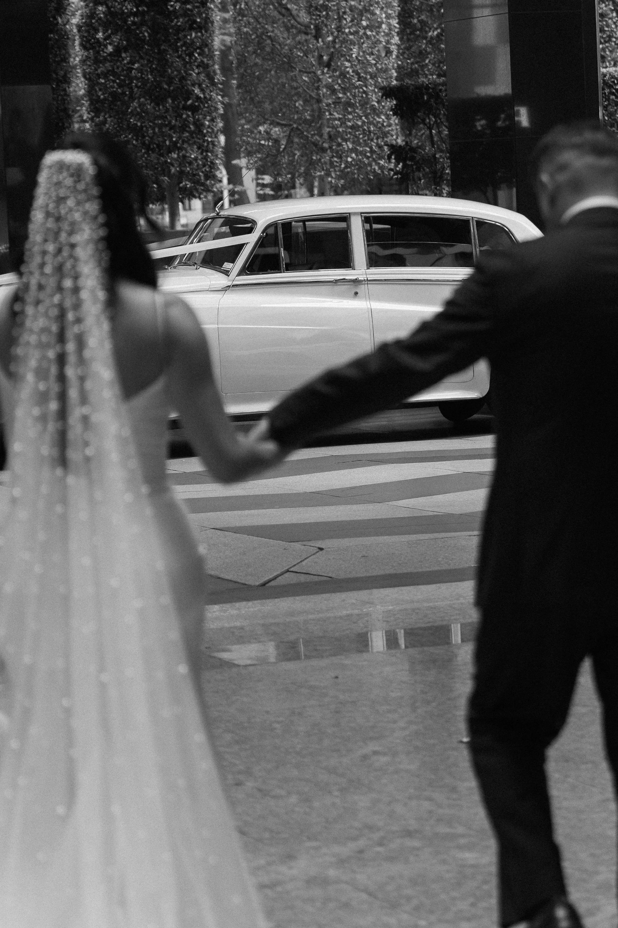A black-and-white photo of a bride and groom holding hands, walking outside near a vintage car, with trees and buildings in the background.