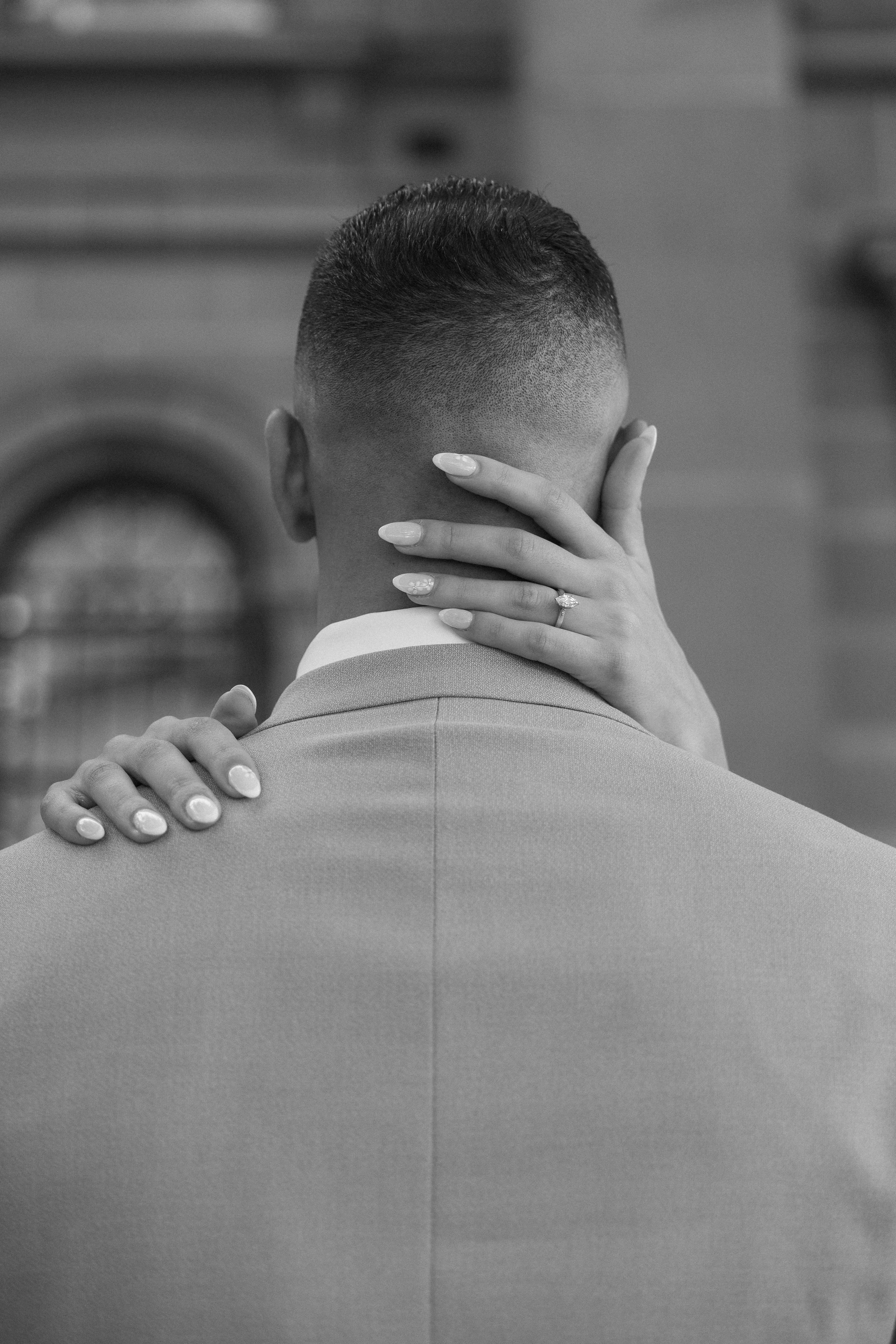 A close-up of a woman with manicured nails, wearing a ring, embracing a man dressed in a suit with a short haircut, with her hand on his shoulder and face hidden behind his head, in an outdoor setting.