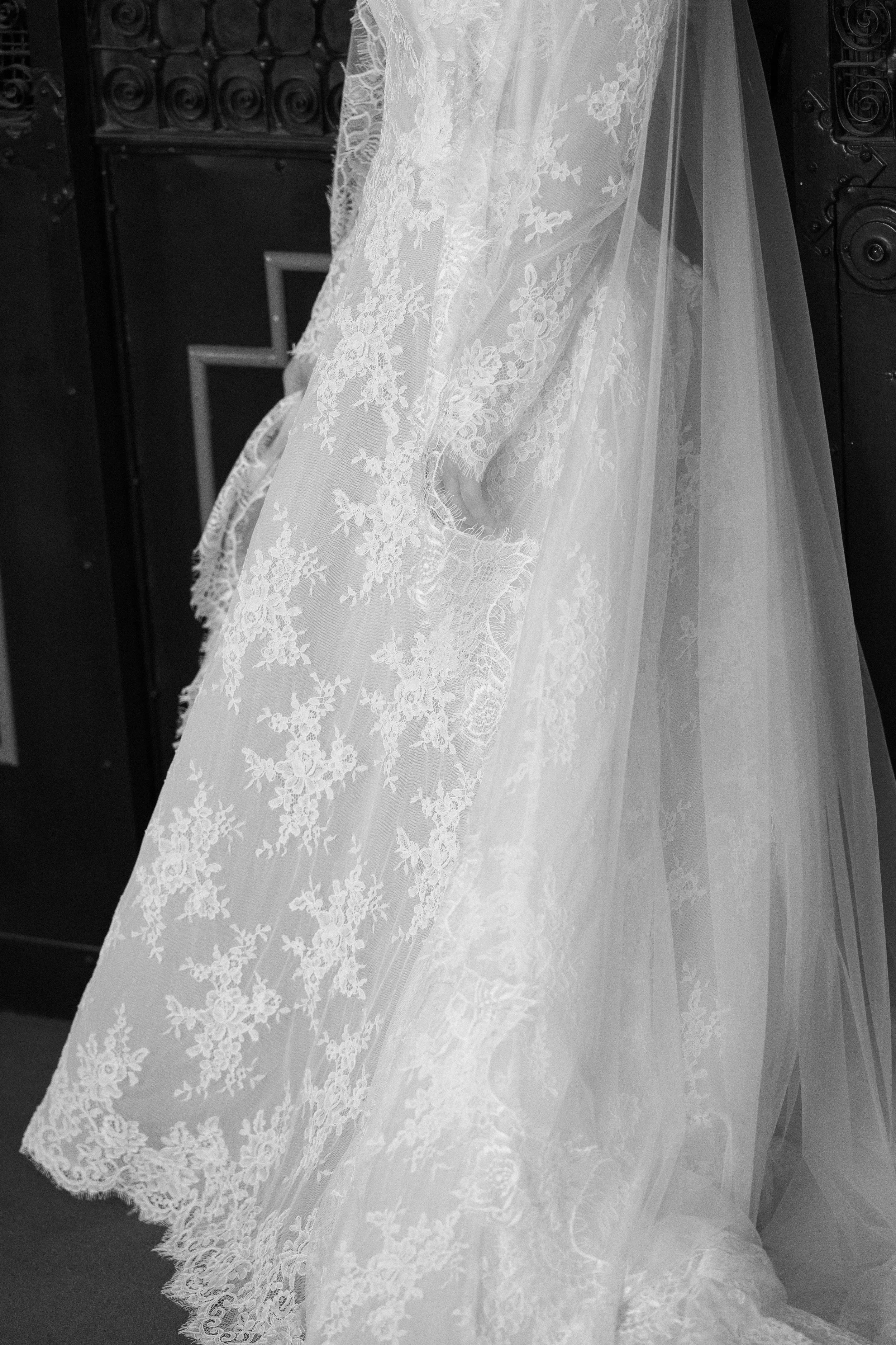 Close-up of a wedding dress with intricate lace and embroidery details, hanging against a dark background.